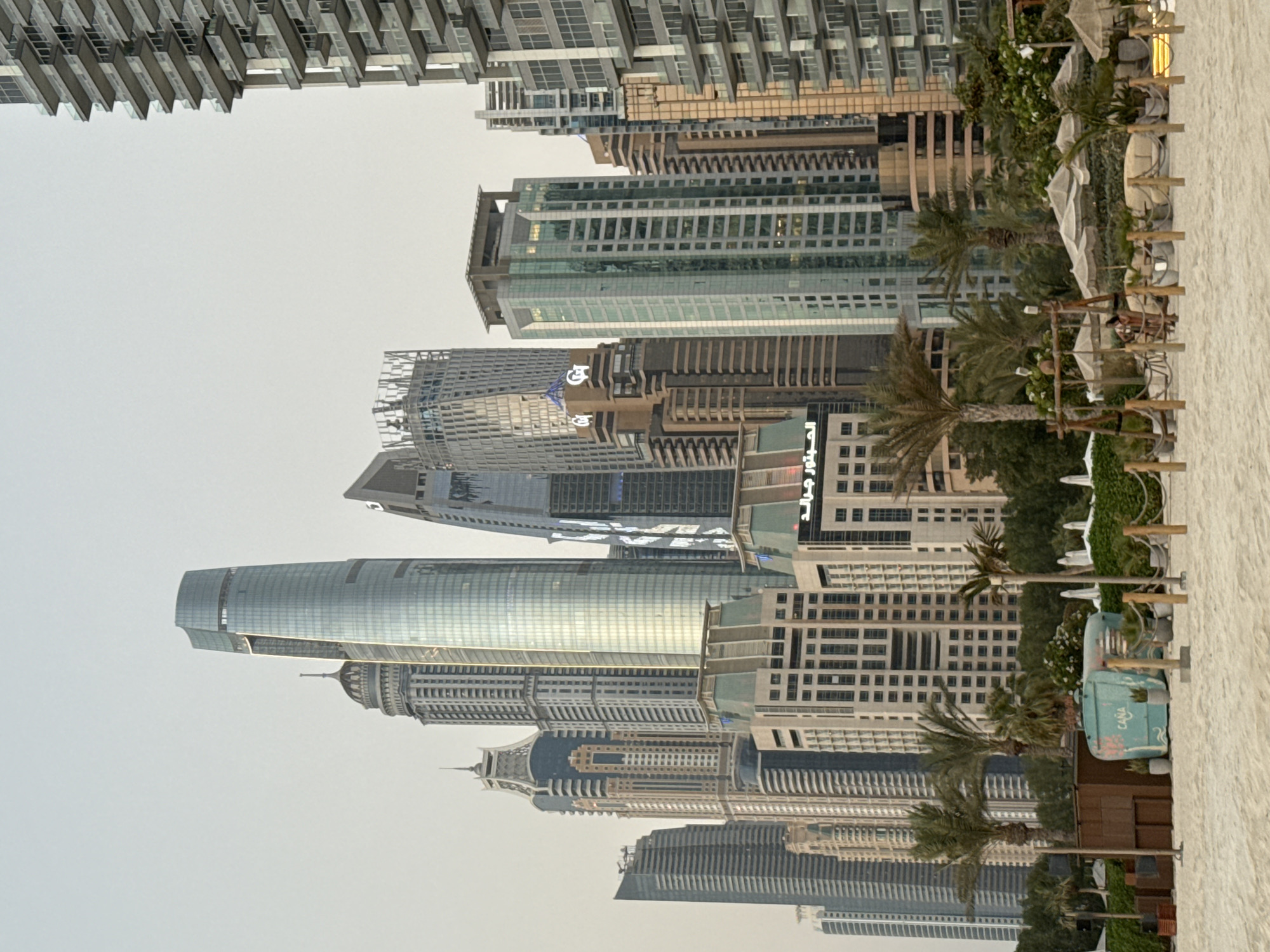 The image depicts a beach scene in Dubai, United Arab Emirates, with a backdrop of modern skyscrapers. The foreground consists of a sandy beach with some beach furniture, including lounge chairs, umbrellas, and a small wooden structure. Palm trees are scattered across the scene, adding to the tropical ambiance. In the mid-ground, a turquoise-colored kiosk with the word "CANA" is visible. The background is dominated by a cluster of impressive high-rise buildings, characteristic of Dubai's impressive urban landscape. The sky is overcast, suggesting it might be early morning or late afternoon.