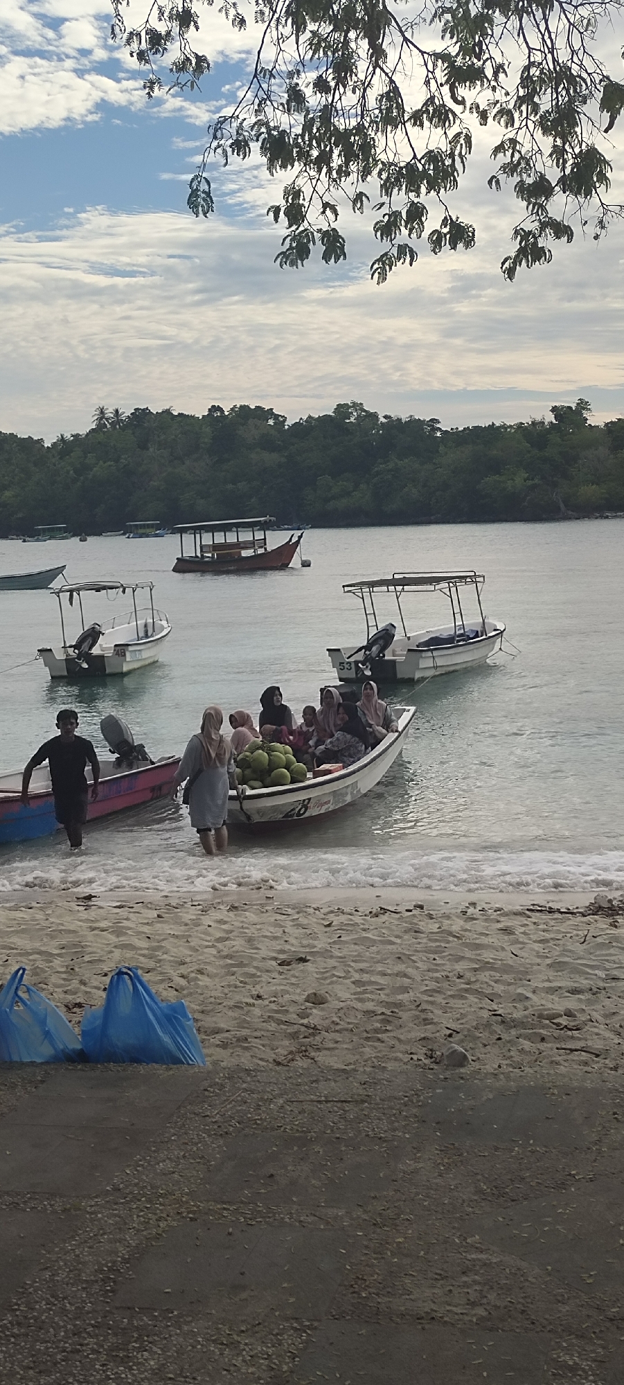 An aerial view from a hill looks down upon a coastline in the city of Sabang, Indonesia. The scene is a beach with several boats in the shallow water. In the foreground, two bright blue plastic bags sit on the sand. Further back, a man in a black shirt is walking in the water towards a pink and blue boat. Several women in hijabs are in a white boat filled with coconuts and what appears to be food items. Other boats are anchored in the water, and a densely wooded island is visible in the background. The sky above is partly cloudy with a pale blue hue.
