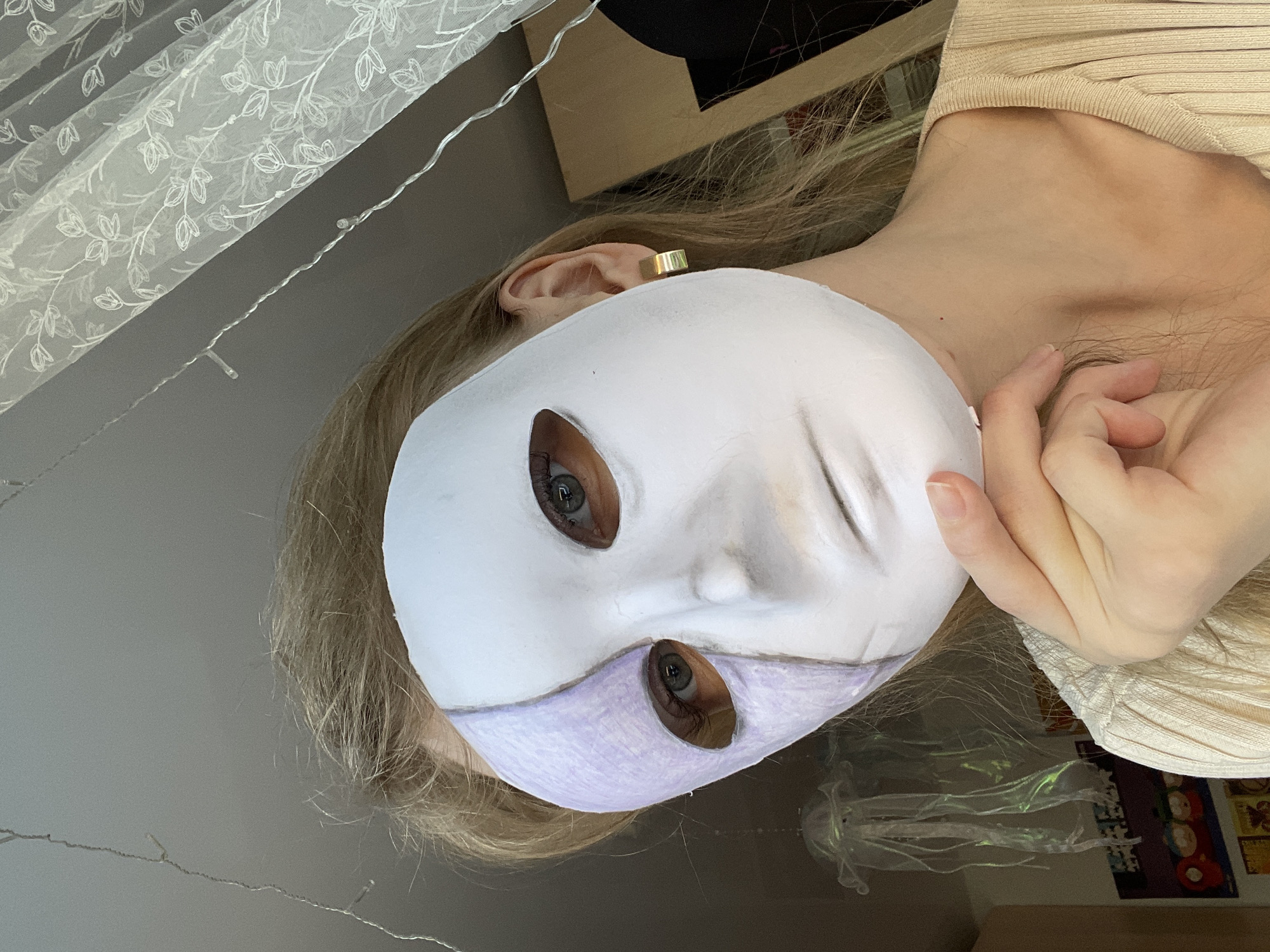 A light-skinned person is holding a white mask with eye holes in front of their face. The lower portion of the mask is colored light purple. The person's blond hair is visible around the mask. The person is wearing a beige top. An earring is visible on their left ear. Background elements include a gray wall, a white curtain with a floral pattern, and string lights. There is a shelf with some objects in the background. The location is not discernible from the image.