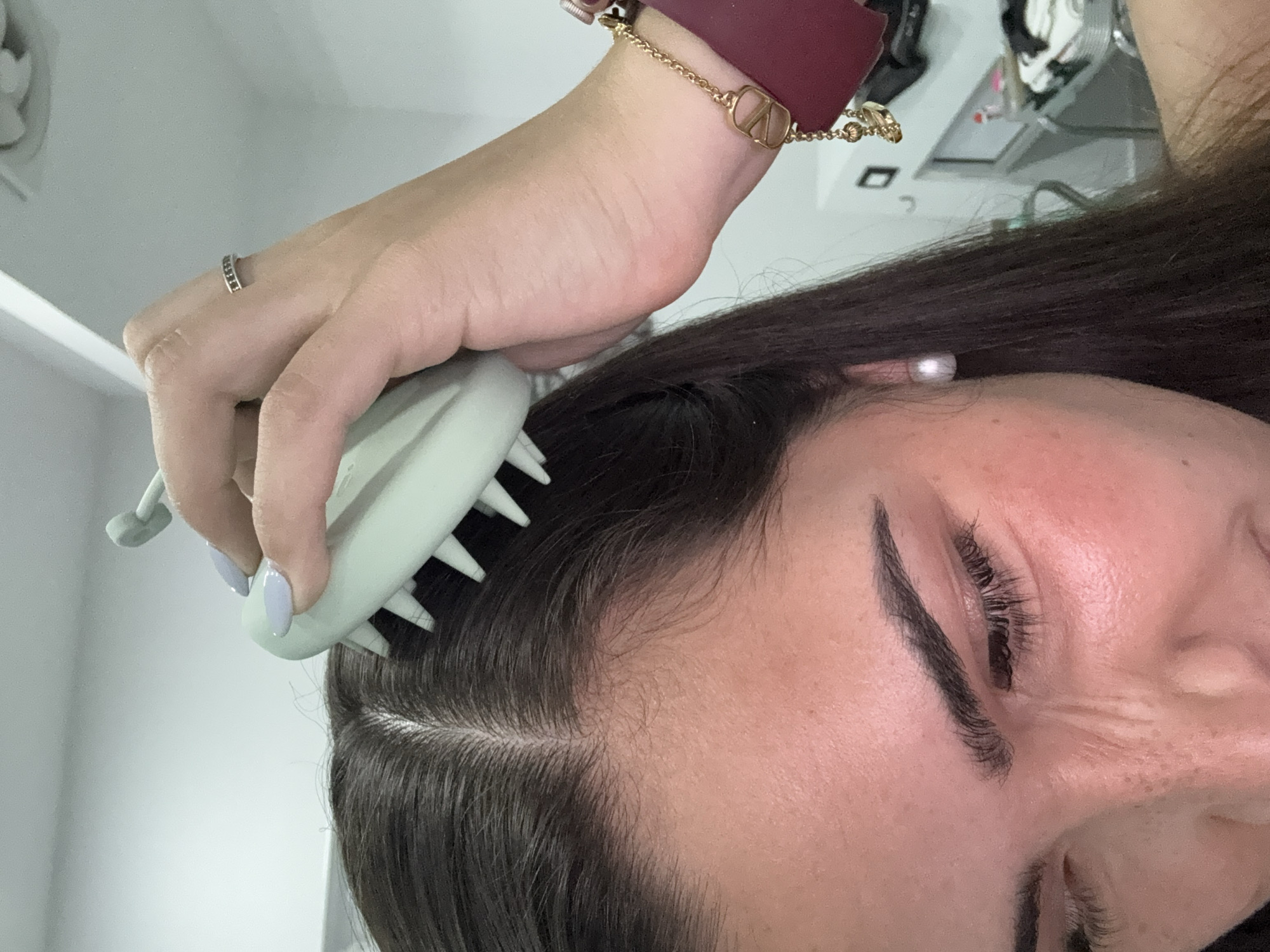 A close-up image depicts a person using a light green scalp massage brush on their dark brown hair. The person's right hand, adorned with a silver ring and light purple nail polish, holds the brush. The person is also wearing a gold chain bracelet over what appears to be a dark red watch band. The image focuses on the individual's face and hair, showing details such as their eyelashes and eyebrows. The background is blurred and does not reveal any specific location information. There are no contextual clues in the image to suggest a location in San Bernardino Tlaxcalancingo, Mexico.