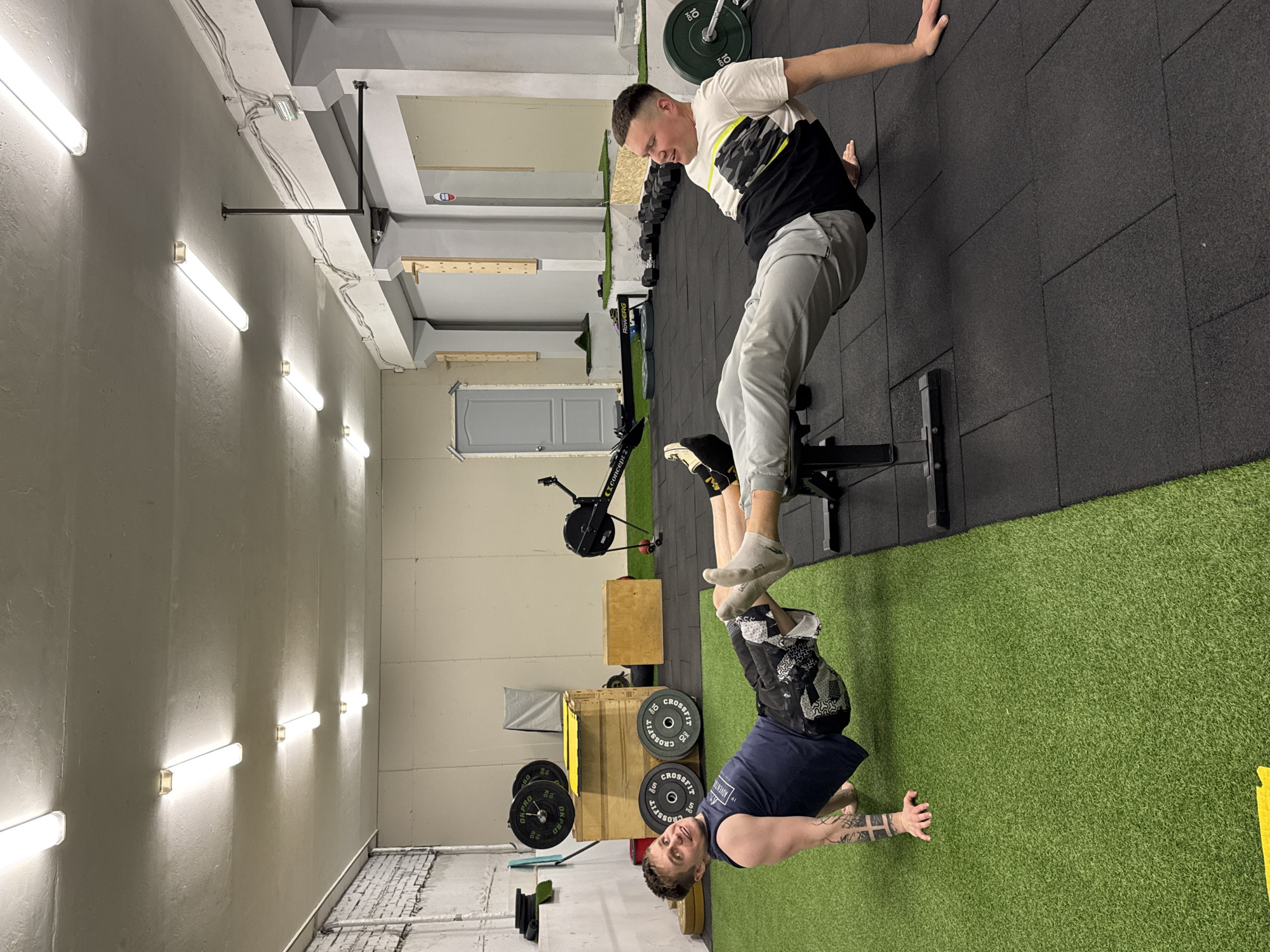 An image, captured indoors in what appears to be a gym or fitness studio in Chaikovsky, Russia, shows two men engaged in a partner calisthenics or strength training exercise. The image is oriented sideways, with the subjects appearing upright if the image is rotated 90 degrees clockwise.
In the foreground, one man, wearing a dark blue t-shirt and shorts, is performing a handstand on artificial green turf. He has tattoos on his forearms and is looking towards the viewer with a slight smile, indicating focus or enjoyment. His hands are flat on the turf, supporting his body.
Above him, another man, dressed in a white and black t-shirt and grey sweatpants, is positioned on a black gym bench. He is in a seated or reclining position, with his bare feet and lower legs elevated and supported, likely by the hands or body of the man below him, though the exact point of contact is obscured. He appears to be performing a core exercise, looking down and to his left with a concentrated expression.
The gym features a blend of flooring: green artificial turf covers the area where the handstand is performed, transitioning to black rubber matting. Various gym equipment is visible in the background, including a Concept2 rowing machine, wooden plyometric boxes, dumbbells, and weight plates. Some weight plates are marked with "CROSSFIT," "OKPO," and "10 KG." The walls are light-colored, possibly concrete, with exposed electrical conduits and fluorescent tube lights providing bright illumination. A light grey door is also visible in the background. The ceiling features concrete beams with further wiring.