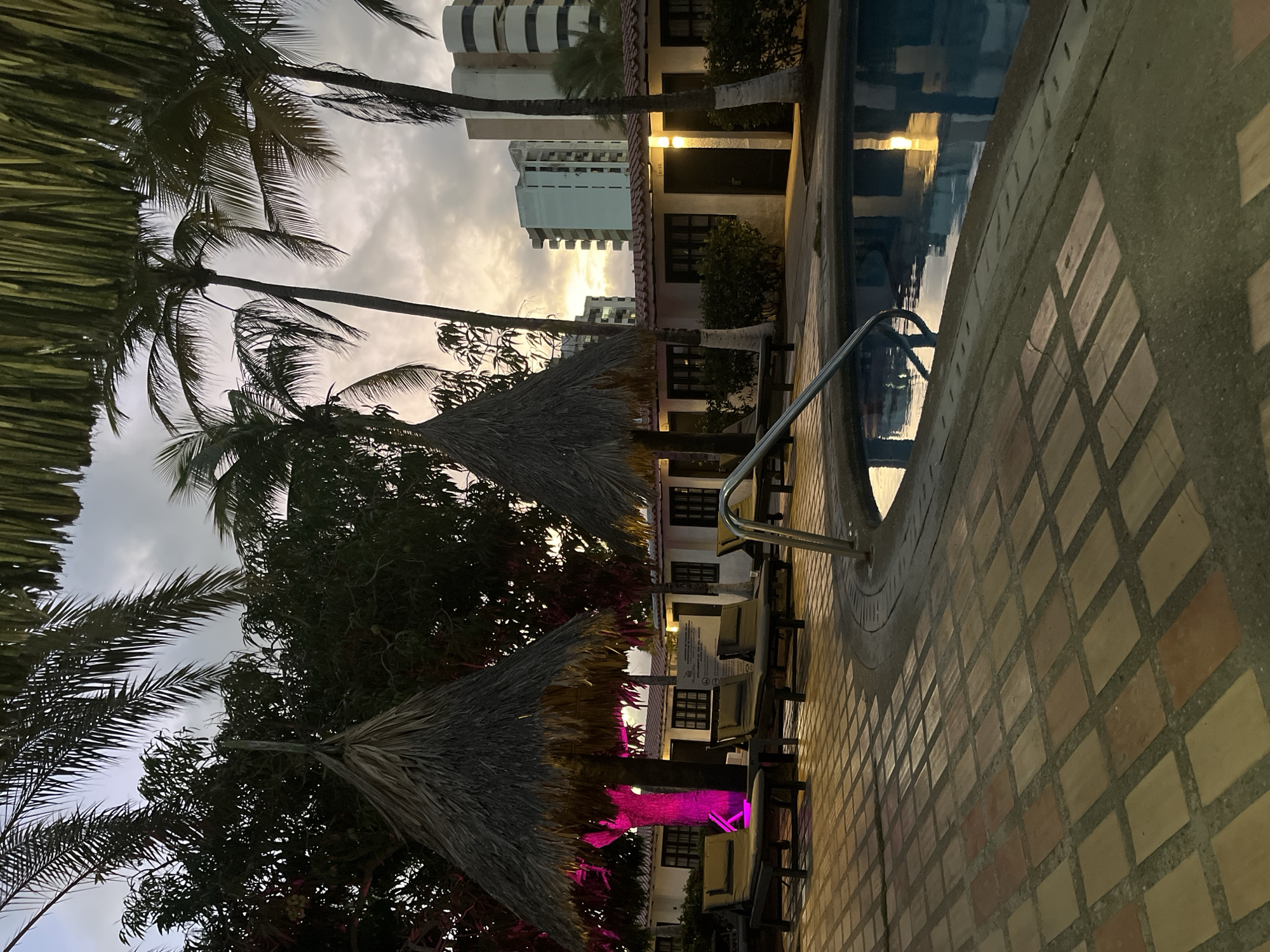 The image depicts a poolside area at dusk in Porlamar, Venezuela. Several palm trees are visible, with one illuminated by pink lighting. Two thatched umbrellas provide shade over lounge chairs. Adjacent to the pool, a series of small buildings with dark windows and white exteriors are lined up, with lighting illuminating some of the doorways. In the background, modern, multi-story buildings rise against a twilight sky with clouds. The ground around the pool is paved with rectangular tiles. The water in the pool reflects the ambient light from the buildings and the sky. There are no people visible in the image.