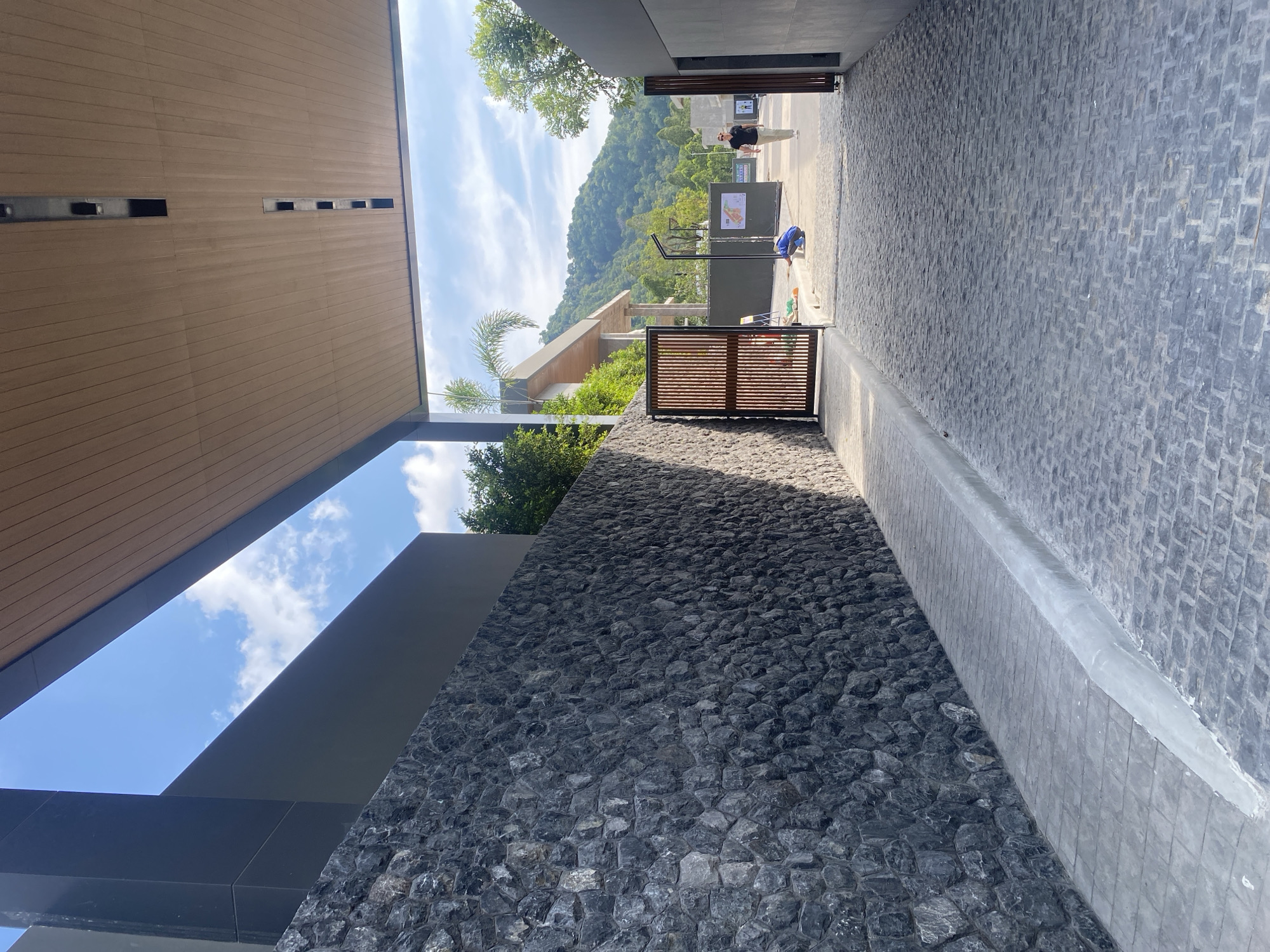 This outdoor shot, taken in Choeng Thale, Thailand, features a stone wall and cobblestone pathway leading away from the viewer. The scene is framed by architectural elements, with a wooden ceiling above and a clear view of mountains and sky in the distance. Two people are visible further down the pathway, one standing and the other appearing to be working on something near the ground. A gate is also present near the wall.