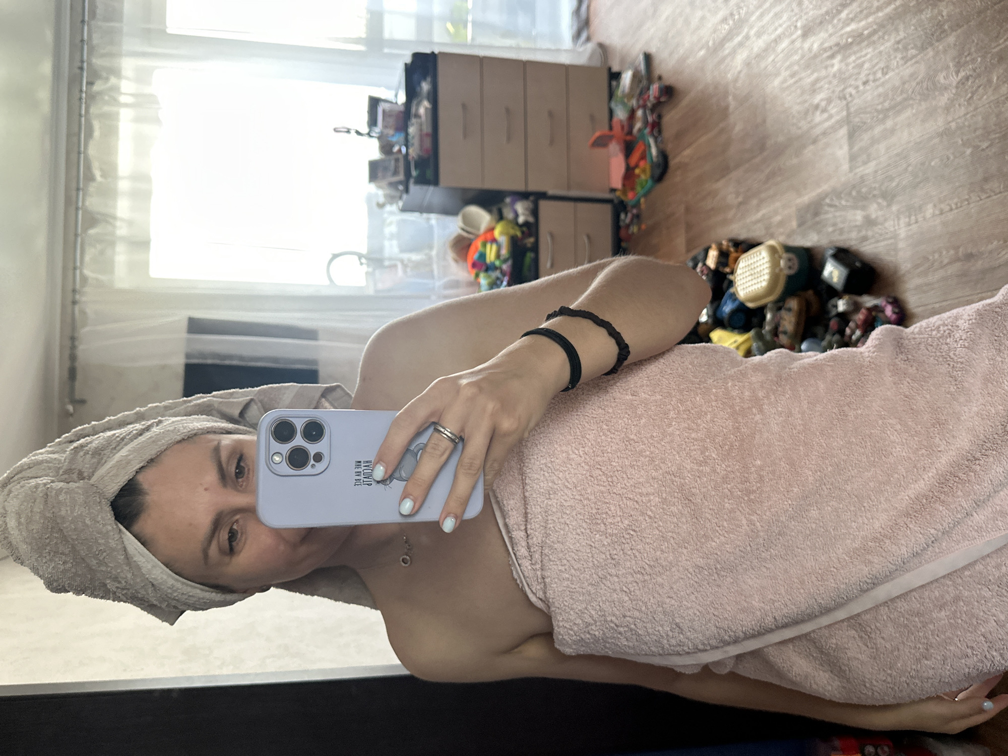 A woman with her hair wrapped in a towel and wearing a pink towel around her body takes a selfie in a room. She is holding a light purple phone in her right hand, which displays a message in Cyrillic and a picture of a bear. The room has a wooden floor and a large window with sheer curtains. To the right, there is a dresser with multiple drawers and a pile of toys on the floor. The woman is wearing a necklace and a ring.