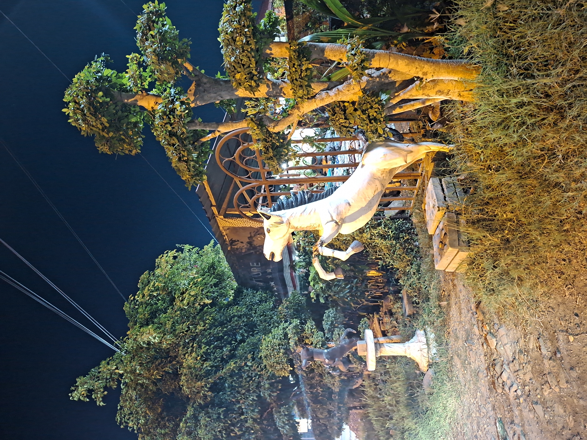 The image shows a roadside display at night, likely in Lhokseumawe, Indonesia. A white horse statue appears to leap over a decorative metal gate. Behind the gate, a tree's branches are illuminated with a warm, orange light. Below the horse, another smaller statue of a donkey is mounted on a white pedestal. Lush greenery surrounds the display. In the upper left of the frame, power lines stretch across a dark blue sky. The ground appears to be dirt or gravel.