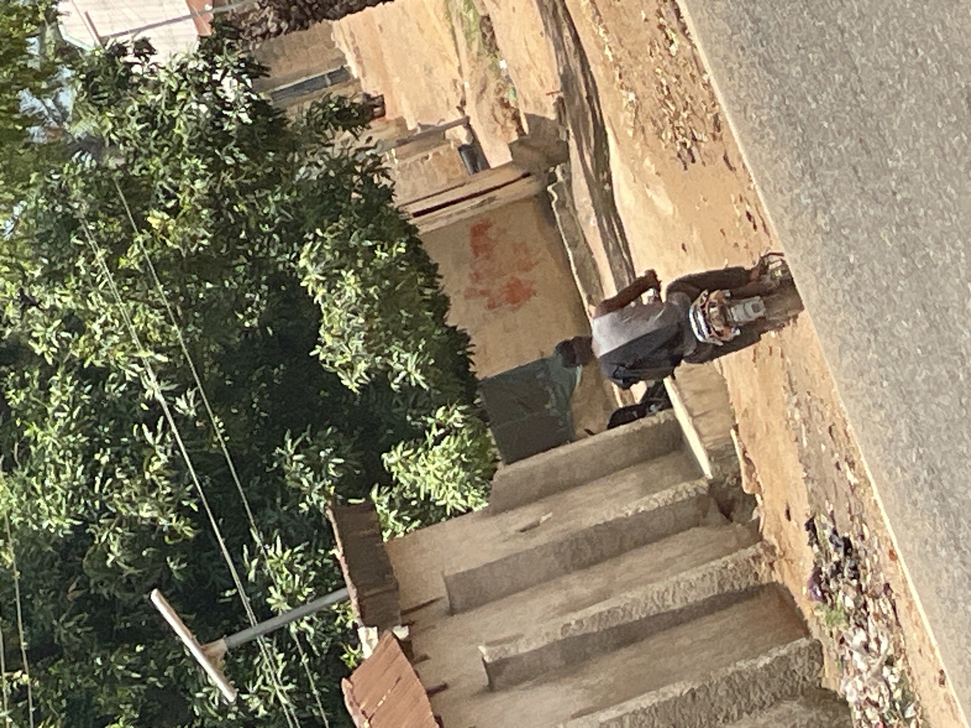 A man walks up a concrete staircase towards a building in Mararaba, Nigeria. A large tree stands to the left of the stairs. The street is to the right of the stairs.