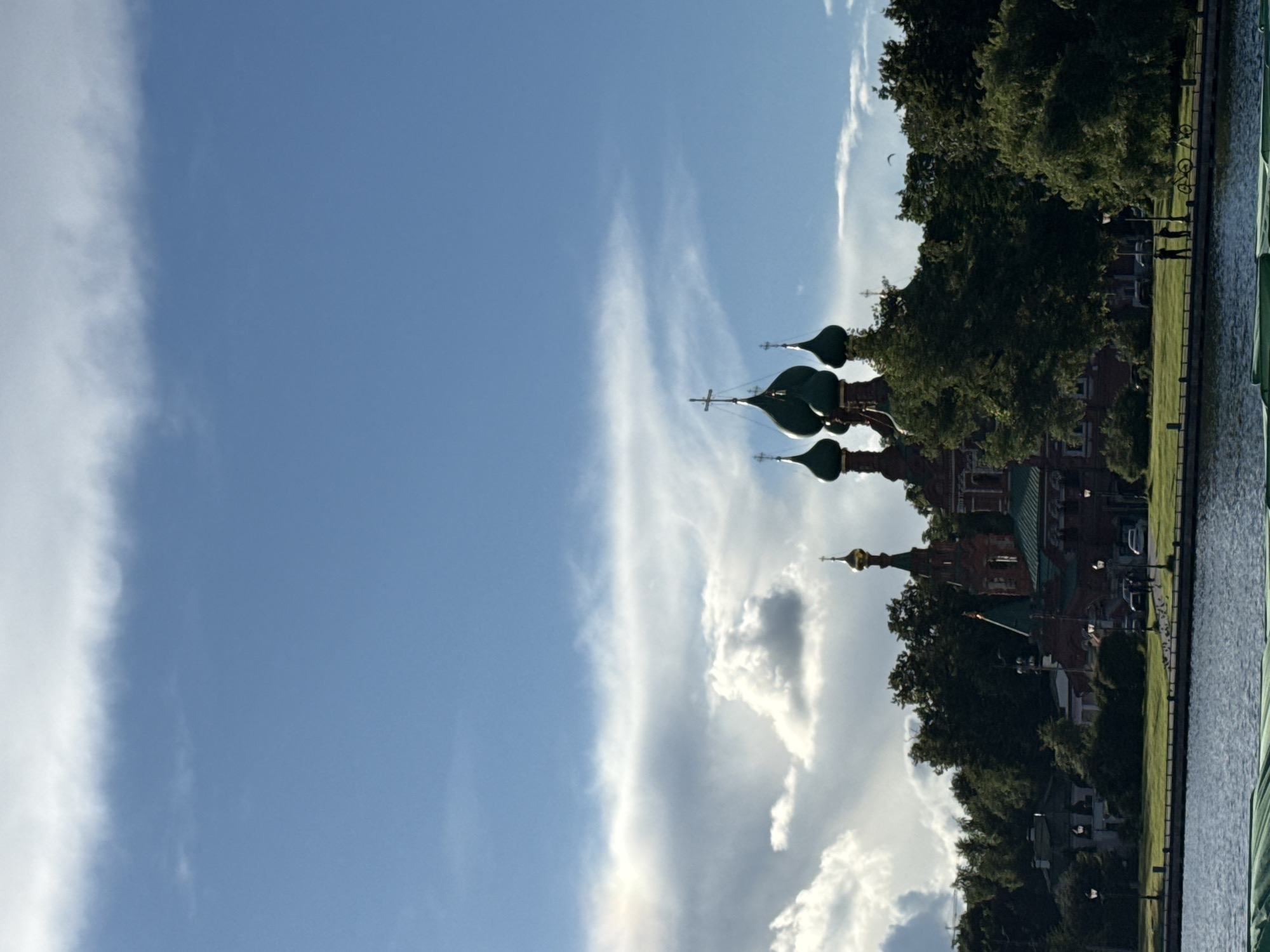 A wide-angle, eye-level shot captures a serene scene of the Church of the Transfiguration of the Saviour in Ostrov, Moscow, Russia, set against a dramatic sky with a mix of bright blue and heavy, white clouds. The church, constructed of red brick with distinctive green domes and gold crosses, stands prominently amidst a dense arrangement of dark green trees. In the foreground, a calm body of water reflects the sky. A paved pathway lines the edge of the water, separating it from a lush green lawn where two figures stand near bicycles. The overall atmosphere suggests a pleasant day, possibly late afternoon, given the lighting.