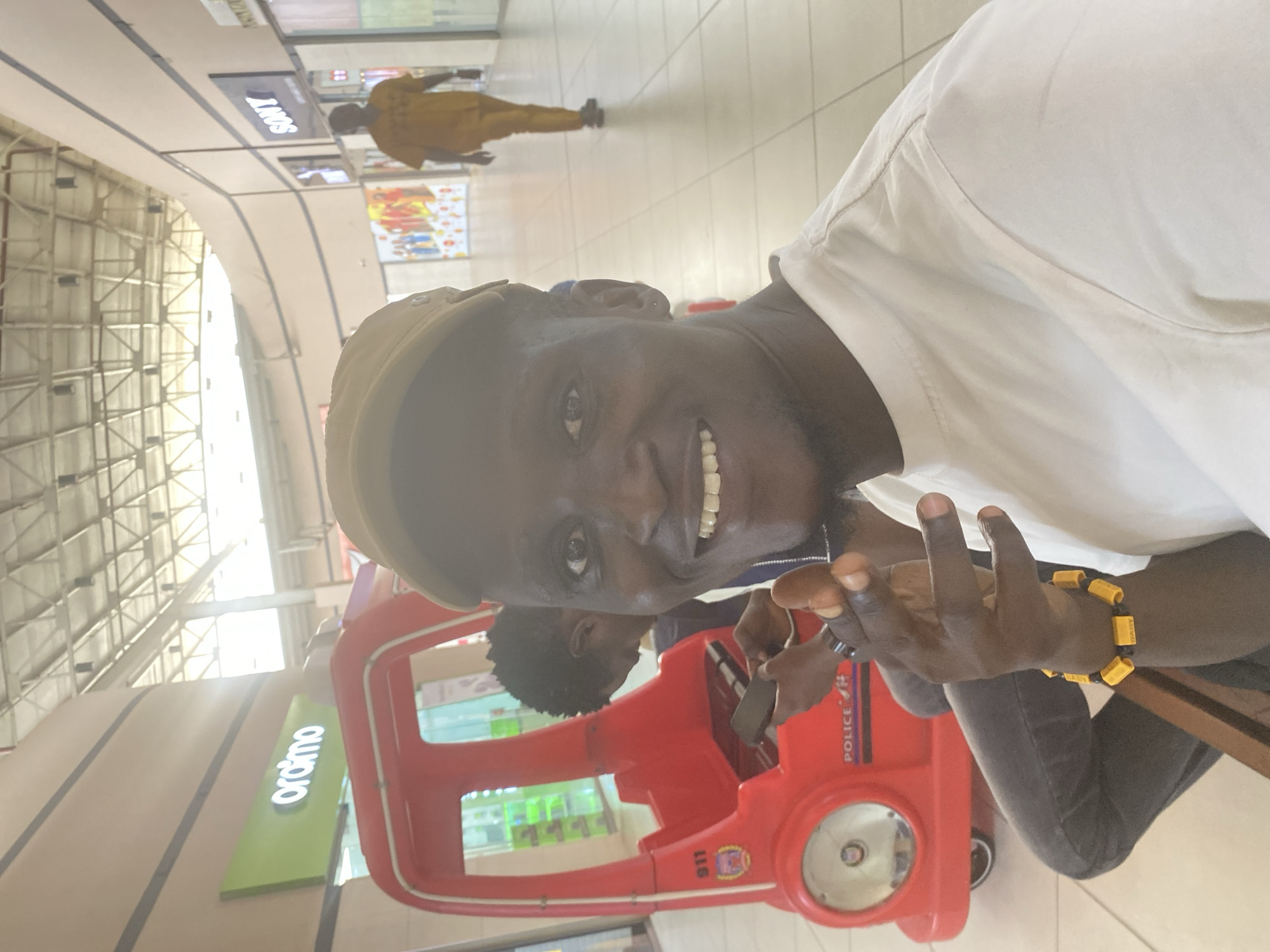 A portrait-oriented photograph, likely taken in a shopping mall in Ikeja, Nigeria, features a smiling Black man as the primary subject. He wears a light-colored (possibly white) shirt and a tan cap, looking directly at the camera with a wide, joyful smile that shows his teeth. A black and yellow beaded bracelet is visible on his right wrist. His hands are positioned in front of him, seemingly clasped or gesturing.
Behind him, another person with dark, curly hair is partially visible, seated in a bright red toy car. The toy car features text such as "POLICE" and "911" on its side, suggesting it's a child's police or fire engine toy. The background reveals an indoor commercial space with a tiled floor and a high, arched ceiling featuring exposed metal framework, through which bright natural light streams.
In the mid-ground, a black sign displaying "SONY" (inverted) is visible, along with a green sign showing "oraimo" (also inverted). Further in the distance, another individual wearing a mustard yellow or light brown uniform is walking away. The scene is well-lit, suggesting daytime, and the overall mood conveyed by the main subject's expression is cheerful.