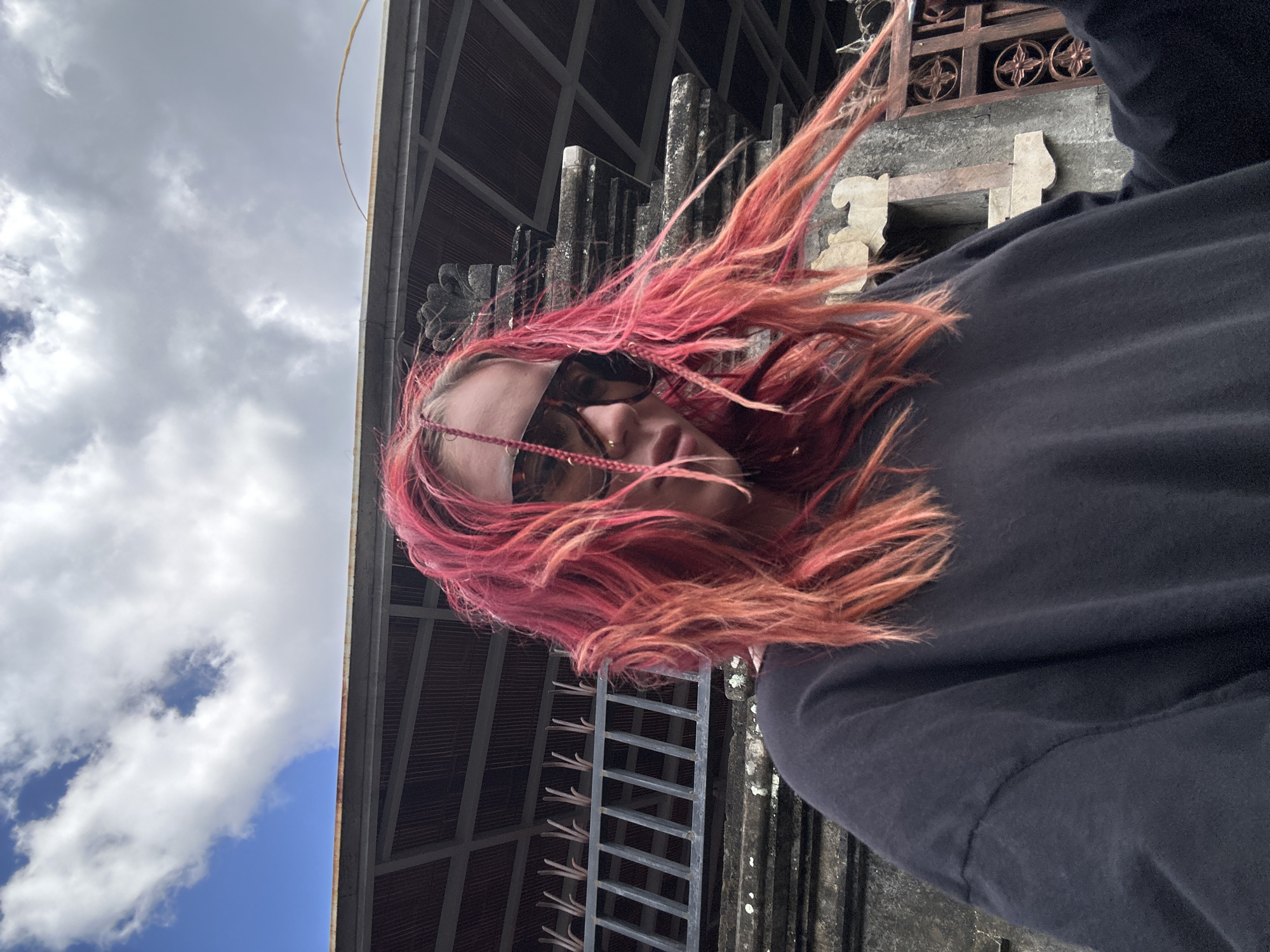 In North Kuta, Indonesia, a young woman with vibrant pink, shoulder-length hair, styled in loose waves and two thin braids framing her face, is captured in a low-angle selfie. She wears large, dark sunglasses and a simple black t-shirt. Her hair is blowing to the right, suggesting a breeze. Behind her, a weathered stone structure with intricate carvings and a dark metal fence with vertical bars is visible. Above the structure, a portion of a dark, sloped roof with visible support beams is present. The sky above is a mix of bright blue and scattered white clouds. The lighting indicates it is daytime.