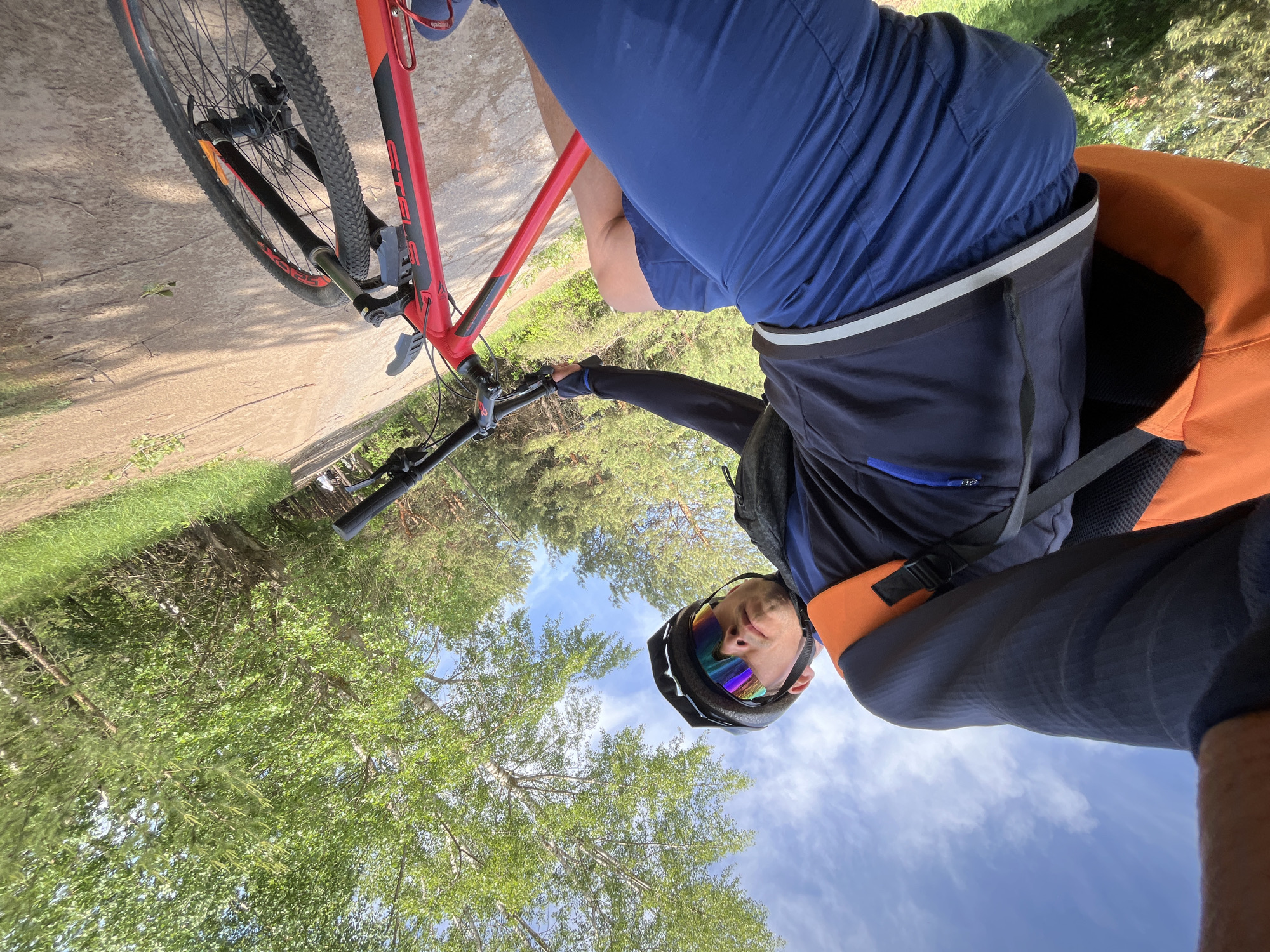 A cyclist wearing a helmet and sunglasses is on a red and black mountain bike. The cyclist is wearing a blue shirt and shorts, along with an orange backpack. The bike is on a paved path with trees and sky visible in the background. The photo was taken from a low angle. Based on the location metadata (Chaikovsky, Russia), the scene is likely set in or around a park or natural area within the city.