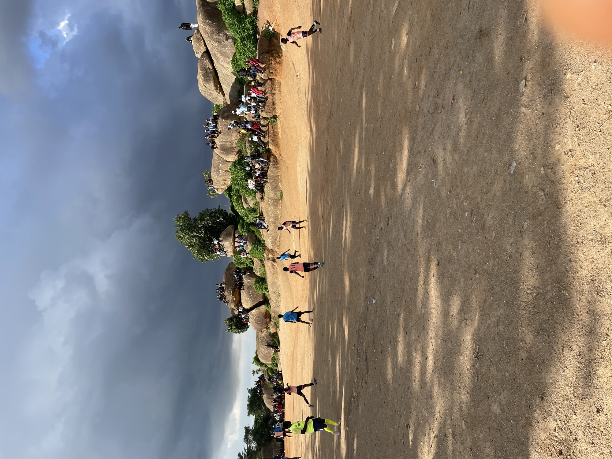 This is an outdoor photograph taken in Jos, Nigeria, depicting a soccer match on a dirt field. Several young men are actively playing soccer, with a ball visible in the center of the field. Spectators are seated and standing on a rocky hillside overlooking the game, some of whom are gathered on large boulders. The sky is overcast with dark, stormy clouds, contrasting with the bright, sunlit area where the game is taking place. Trees and greenery are present on the hillside, interspersed with the rocks. The ground is a dusty, reddish-brown earth, with some scattered debris. The overall scene suggests a local sporting event with a substantial audience participating in the atmosphere.