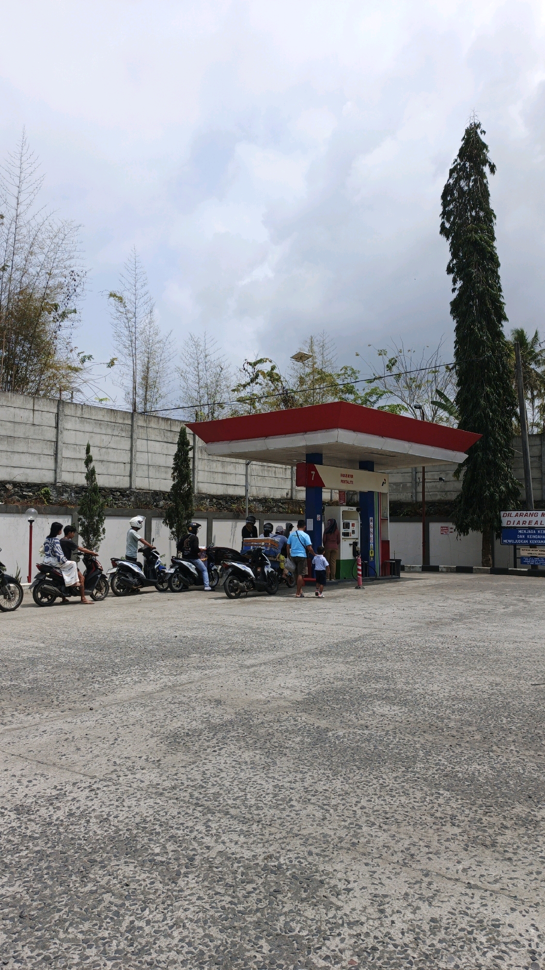 In Sukaresik, Indonesia, a busy gas station has several people on motorbikes refueling. A red and white canopy covers the pumps. A tall, thin evergreen tree stands to the right of the station, and a concrete wall runs along the background, with bamboo and other vegetation visible behind it. The sky is overcast.