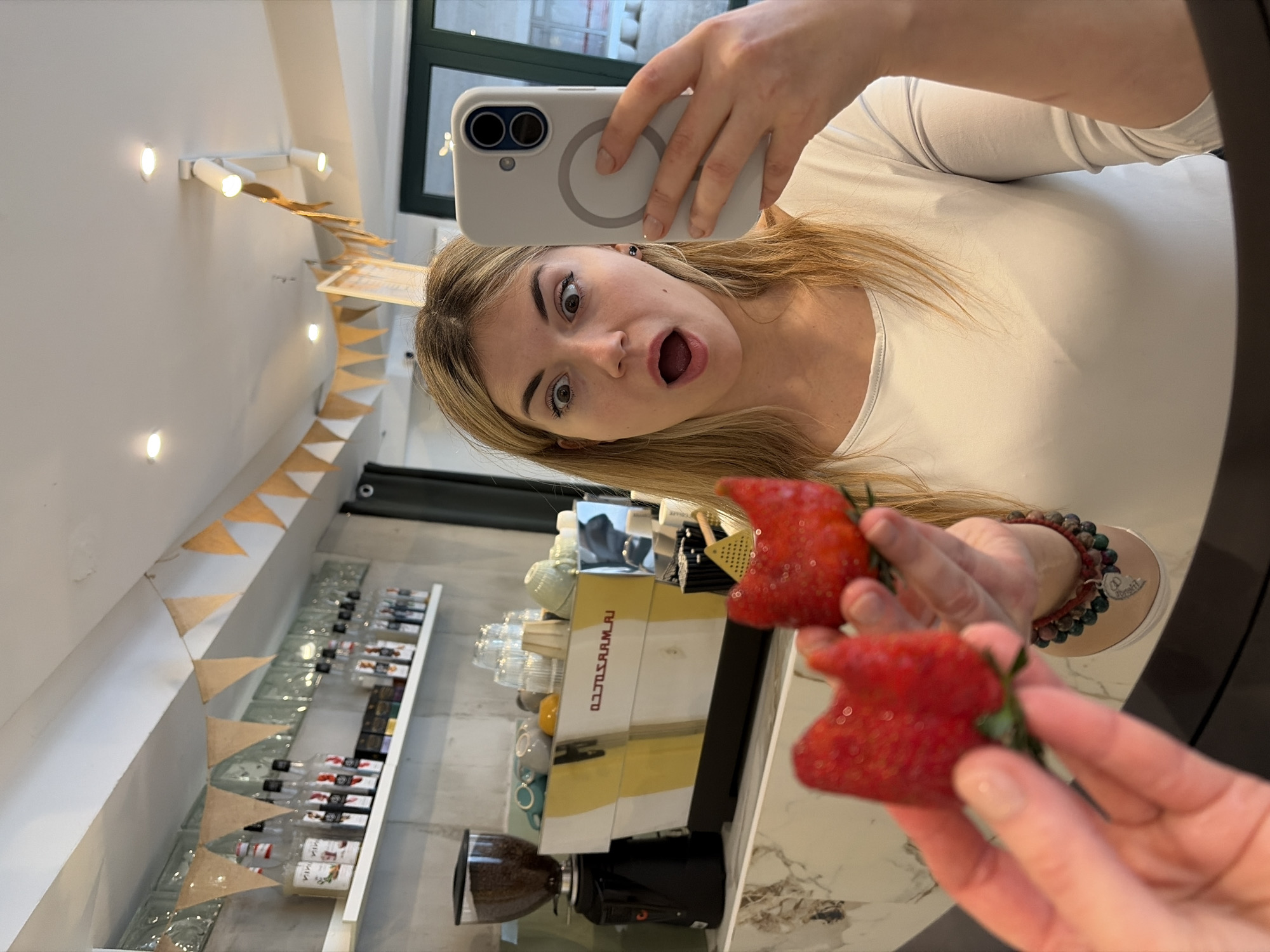 A woman with blond hair is taking a selfie while holding two strawberries. She is wearing a white long-sleeved shirt and a bracelet with beads. The background appears to be a cafe or bar with a shelf of bottles and a coffee machine. There are also party decorations on the wall. Based on the style and decor, it is possible that this is taken in a modern cafe in Budva, Montenegro, but the image alone does not confirm the location.