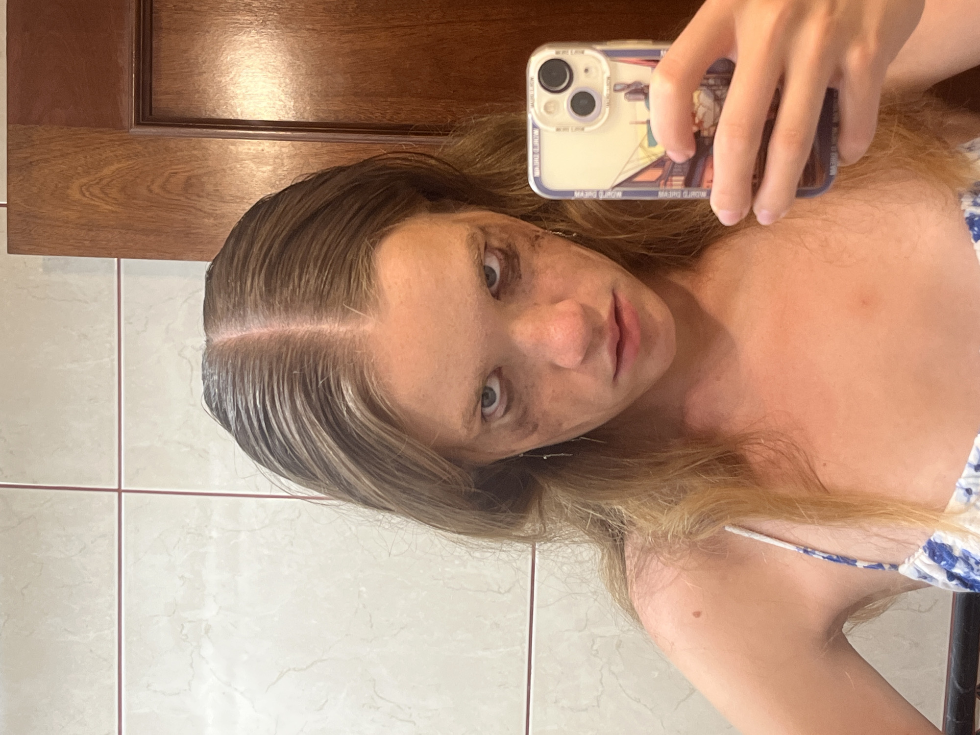The image is a selfie taken by a young person in a bathroom. The person is holding a smartphone, which is angled to capture their reflection in a mirror. The person has light brown hair and is wearing a blue and white patterned top. Their face is turned towards the camera, and they appear to be looking directly at it. Some smudges of dark color are visible on their face, particularly around the eyes and cheekbones, which might suggest makeup or dirt. The background consists of tiled walls and a wooden cabinet or door. The lighting appears to be artificial, as is common in indoor settings. The overall impression is that of a casual, personal moment. The image does not contain any overt signs of a specific time of day or weather conditions.