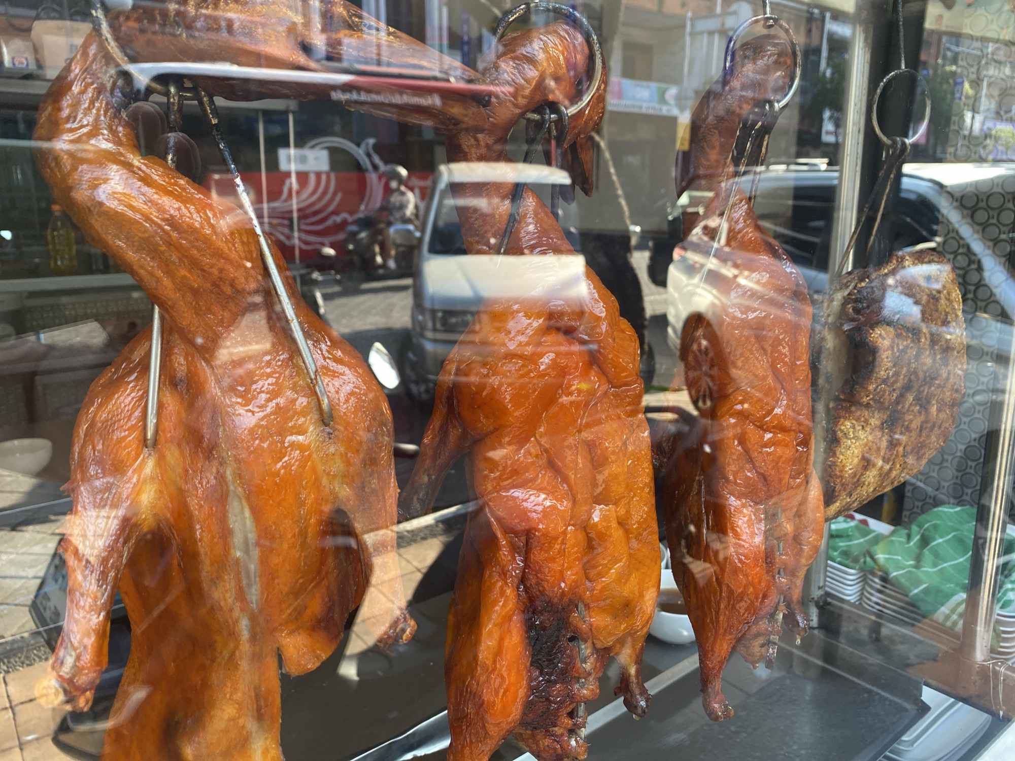 A row of whole roasted ducks hang from metal hooks and rods inside a glass display window, likely a restaurant or market in Denpasar, Indonesia. The ducks have a glossy, golden-brown skin, indicating they are freshly roasted. The display is illuminated by natural sunlight, creating reflections on the glass. In the background, through the display window, a street scene is visible with a white van, a person on a motorcycle, and other buildings. Inside the display, stacked white bowls and a green and white striped cloth can be seen.