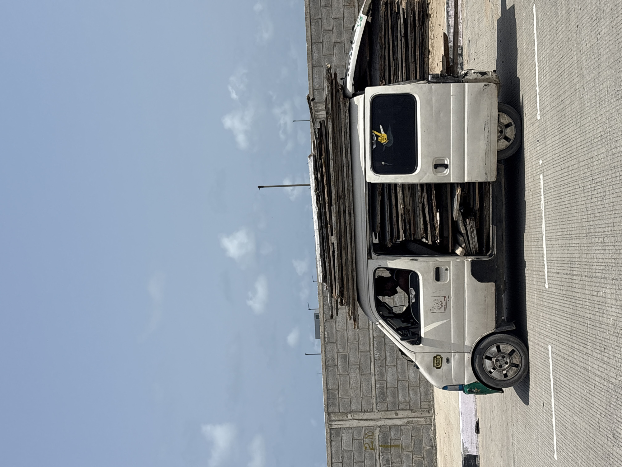A white utility van, heavily laden with long, dark-colored materials resembling wooden planks or panels, is depicted parked or stopped under a bright, clear blue sky with a few scattered white clouds. The image is taken in daylight, suggesting sunny weather.
The van has its front passenger door and the rear side door on the visible side open. The cargo extends high above the van's roof and fills the interior. A person with dark hair is visible in the driver's seat. On the rear side window, a distinctive yellow and black decal, possibly a cartoon character, is affixed. Another circular emblem or logo is visible on the driver's side door. Near the front wheel, a small box with the letters "HDF" is visible on the side of the vehicle, and a green and white object, possibly a plastic bottle, rests on the ground.
The van is situated alongside a light-colored concrete block wall, on which the yellow characters "2D" are painted. To the right of the van is a concrete road surface marked with white lines, likely lane divisions. The scene captures a moment of transport or delivery activity, with the van's substantial load highlighting its utility in a bustling environment.