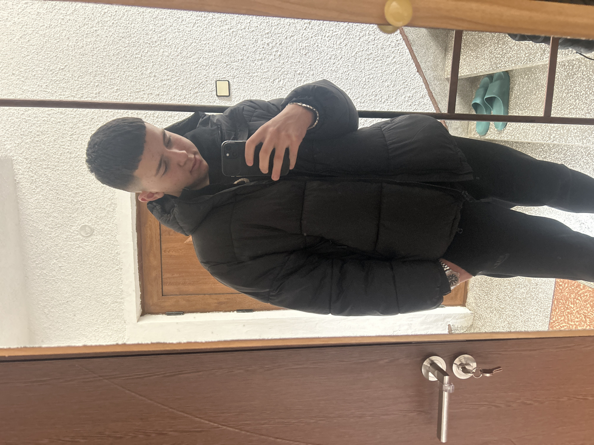 The image is a selfie taken in a mirror indoors. A young man with short, dark hair is holding a smartphone, presumably to take the picture. He is wearing a black puffer jacket and black pants. He also appears to be wearing a bracelet on one wrist and a watch on the other. Reflected in the mirror is a doorway, a textured white wall, a set of stairs with a metal railing, and a pair of blue sandals on one of the steps. The background seems to be an interior hallway or landing. There is a brown door with silver hardware (handle and keyhole) in the lower foreground, also reflected in the mirror. Without additional information, it is impossible to verify the location in the city of Zlatograd, Bulgaria.