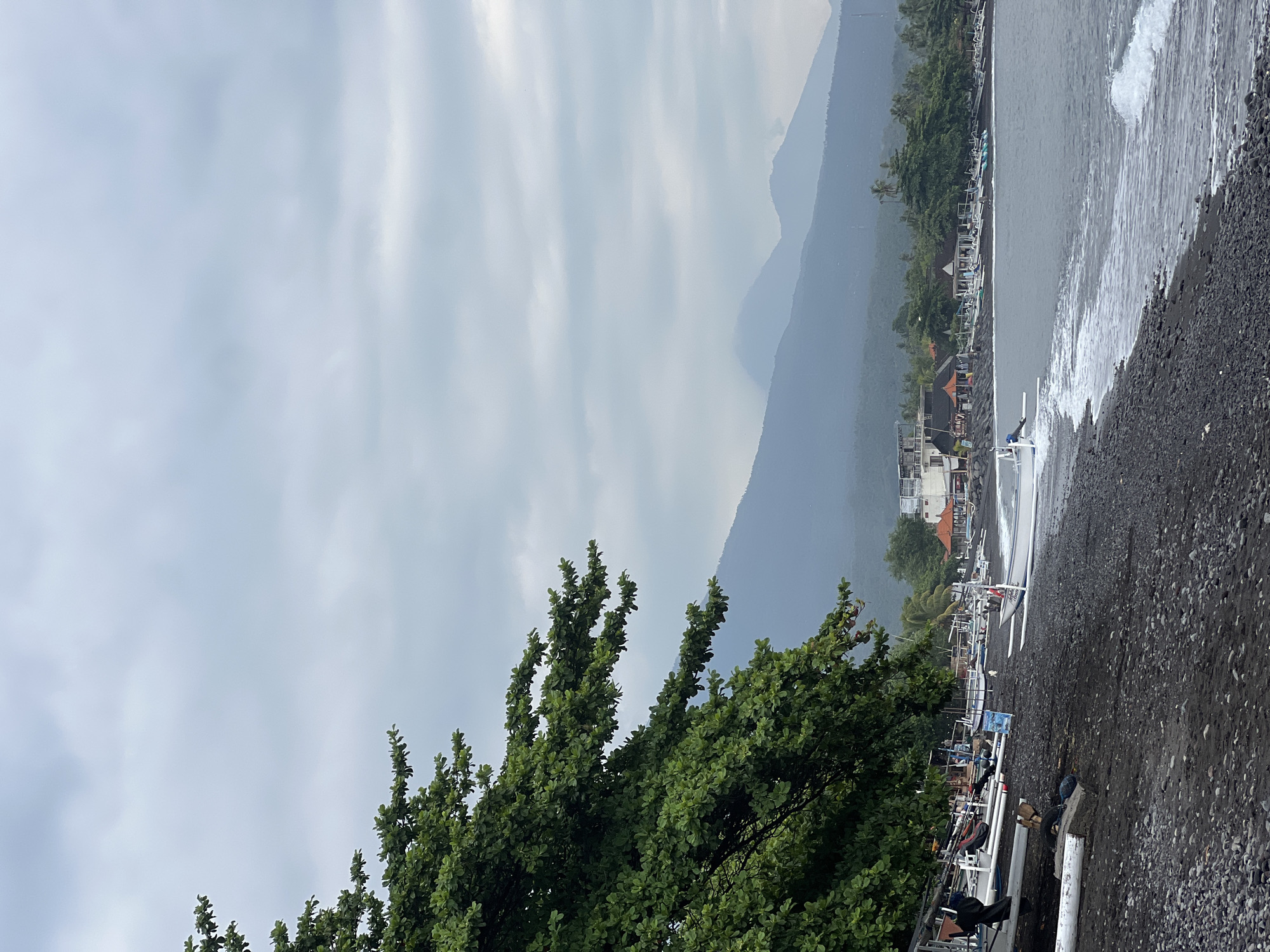 The image depicts a serene beach scene in Abang, Indonesia. The foreground is dominated by a dark, pebble-strewn beach with gentle waves lapping at the shore. Numerous traditional outrigger boats are neatly lined up on the sand, suggesting a fishing village or coastal community. Lush green foliage from a large tree frames the left side of the composition, its branches reaching towards the sky. In the mid-ground, a cluster of buildings, including some with thatched roofs, are visible along the coastline, interspersed with palm trees. The background is characterized by a majestic mountain range, its peaks shrouded in a soft, hazy mist, indicative of a cloudy or overcast day. The overall atmosphere is tranquil and natural.