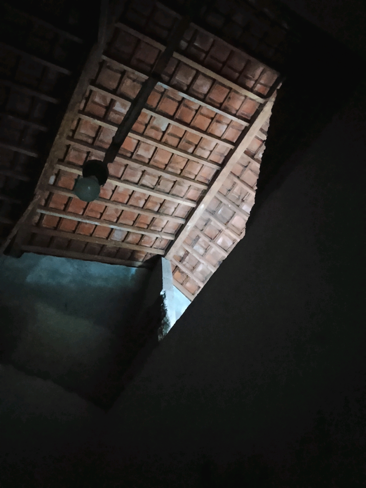 An interior shot looks up at a rustic, exposed roof made of wooden beams and reddish-brown tiles. The tiles are laid out in a grid pattern, with some gaps visible. A single light bulb hangs from the ceiling near the center of the frame. To the left and bottom of the image, dark, unpainted walls and a portion of what appears to be a rough concrete surface are visible. The scene is dimly lit, with shadows obscuring much of the detail. The location is identified as Ujungrusi, Indonesia.