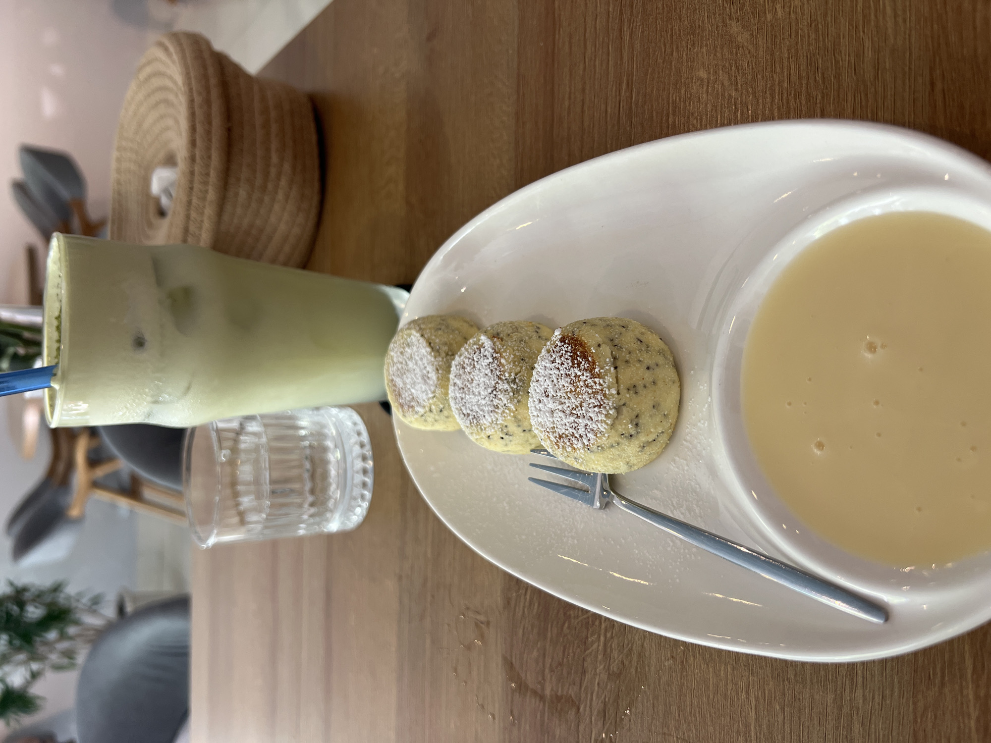 A top-down, close-up shot shows a dessert plate on a wooden table next to a tall glass of a green drink and a small glass of water. The plate holds three small, round cakes sprinkled with powdered sugar, alongside a small bowl of light yellow sauce. A fork rests on the plate, between the cakes and the bowl of sauce. A woven basket is visible in the background. The image was taken in a café in Bắc Nha Trang, Vietnam.