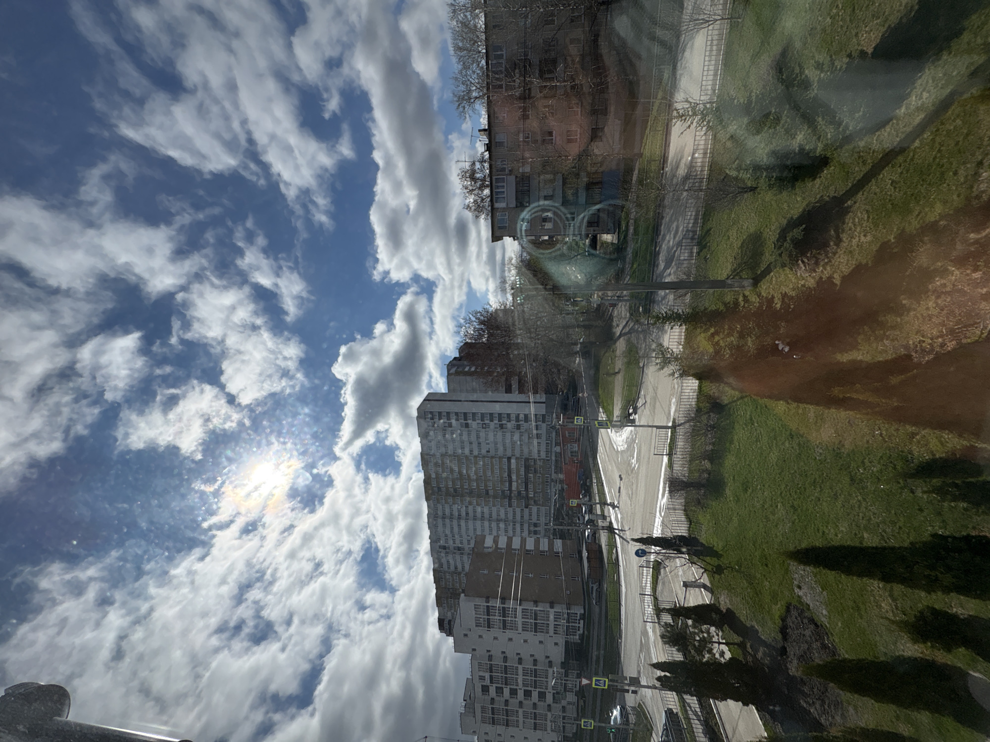 The image shows an urban scene in Ulyanovsk, Russia, possibly viewed through a window. The sky is partly cloudy with bright sunlight. The view includes residential buildings, a street intersection, and grassy areas. The reflection in the window is of the buildings and sky outside.