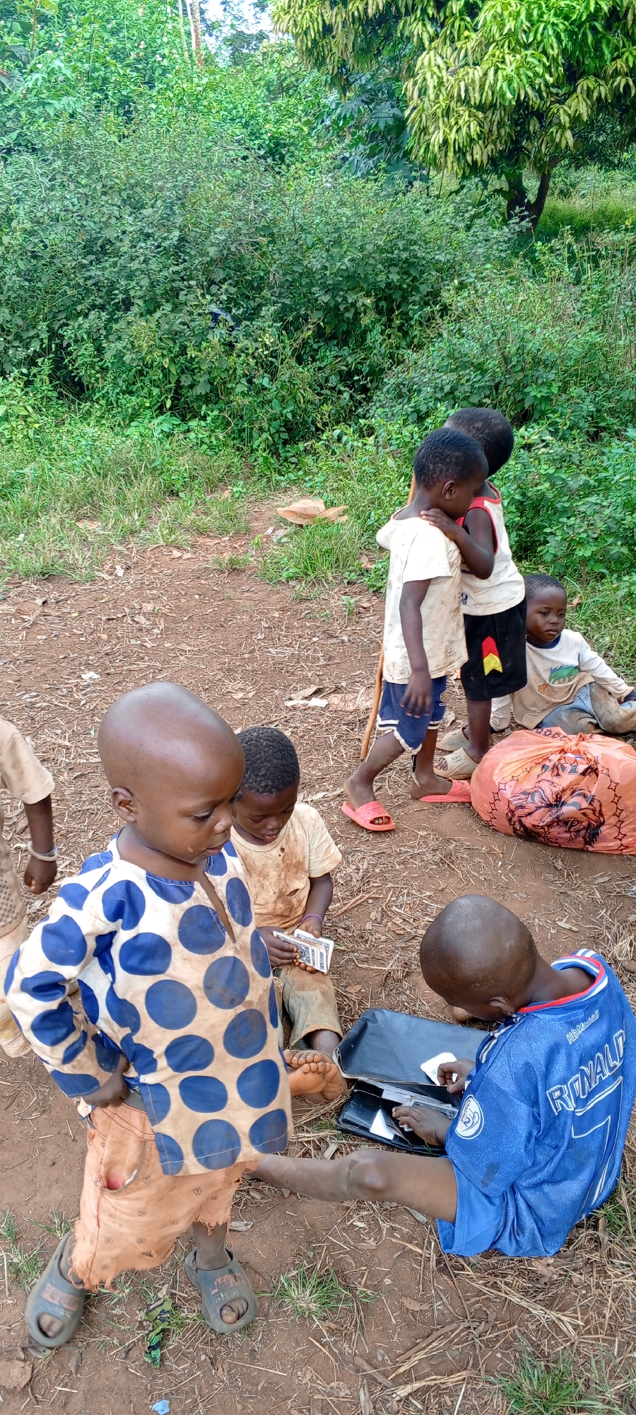 The image shows a group of at least seven young children, primarily boys, gathered outdoors in Ngalula, Democratic Republic of the Congo, during bright daytime. The setting is a dirt clearing with patches of sparse green grass, surrounded by dense green bush and trees in the background, indicative of a rural or natural environment.
In the foreground, a child wearing a blue and white polka-dotted tunic and worn orange shorts stands, looking downwards. To their right, another child sits on the ground, intently counting or examining several banknotes in their hands. Beside them, a third child wearing a blue soccer jersey with "RONALD 7" and "HHB Madrid" printed on it is seated, focused on a black folder or case in their lap.
Other children are visible in the mid-ground. Two children stand closely together, one in a white t-shirt and blue shorts holding a stick, the other in a light top. Another child is seated further back near a large orange plastic bag featuring a dark graphic design. Most children are barefoot or wear simple sandals. Their clothing appears worn and somewhat dirty, consistent with outdoor play. The children's expressions are generally calm and focused on their individual activities or observing those around them. The ground is composed of reddish-brown earth mixed with dry leaves and small debris.