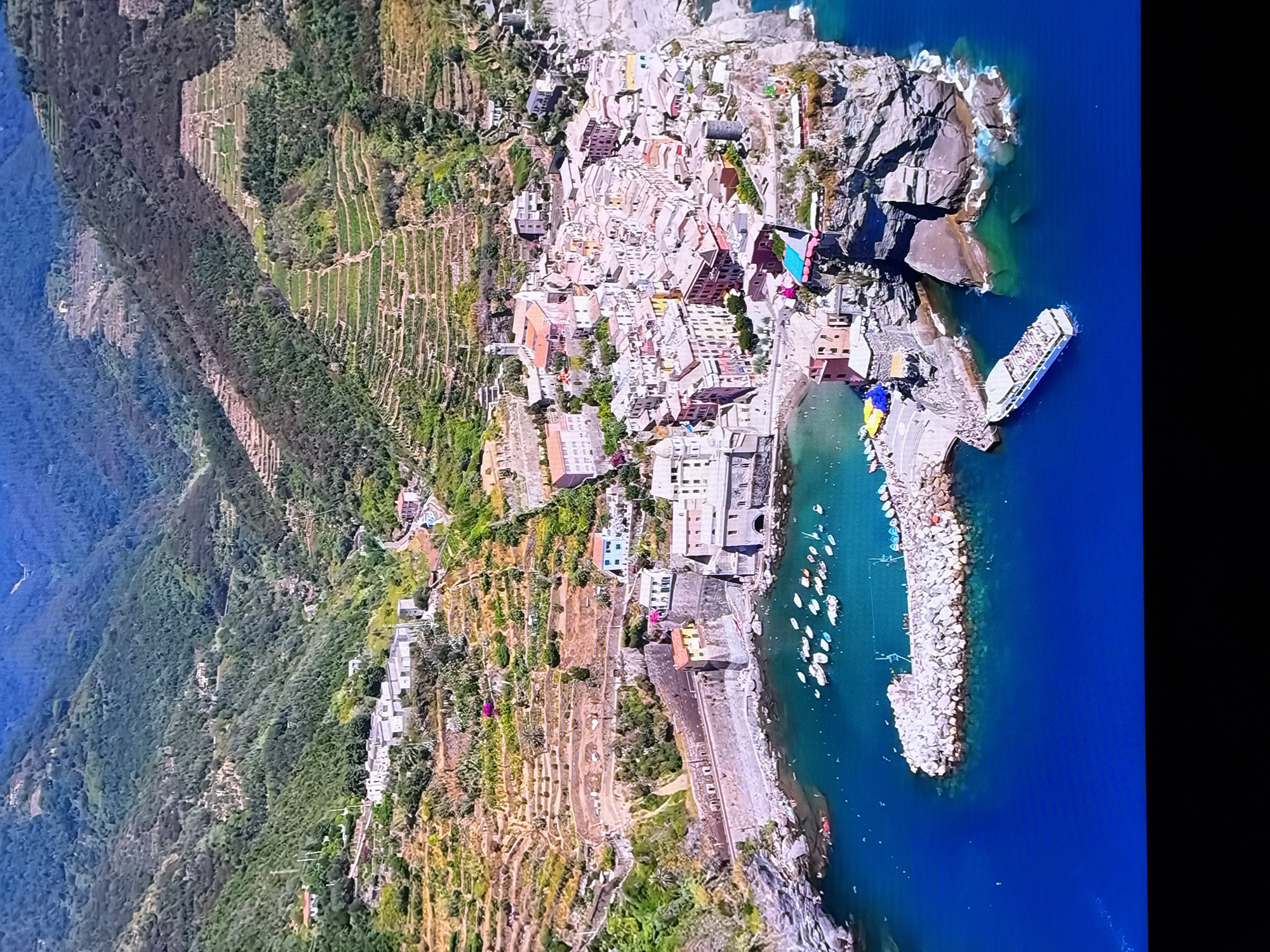 This image is an aerial view of the coastal town of Vernazza, one of the five villages of Cinque Terre, located in the Liguria region of Italy. The town is built on a rocky promontory jutting into the Ligurian Sea. Colorful buildings with red-tiled roofs are clustered tightly together, climbing up the steep hillsides. Terraced vineyards and olive groves are visible on the surrounding slopes. A harbor with numerous small boats is situated in front of the town, protected by a breakwater. A larger passenger ferry is docked to the right of the breakwater. The sea is a vibrant turquoise and blue, with waves crashing against the rocky coastline. The image captures the picturesque and rugged beauty of the Italian Riviera. This location is not in Chirchik, Uzbekistan.