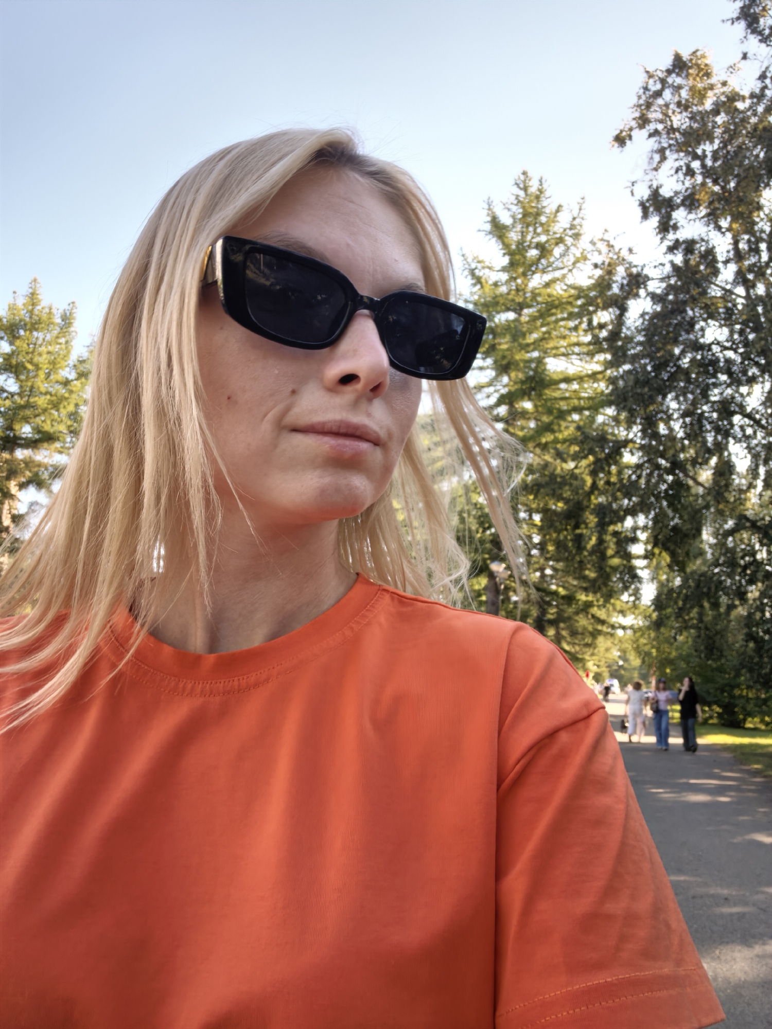 A woman wearing sunglasses and an orange t-shirt stands in a park with trees and a path. In the background, several people are walking on the path. The scene is outdoors on a sunny day.