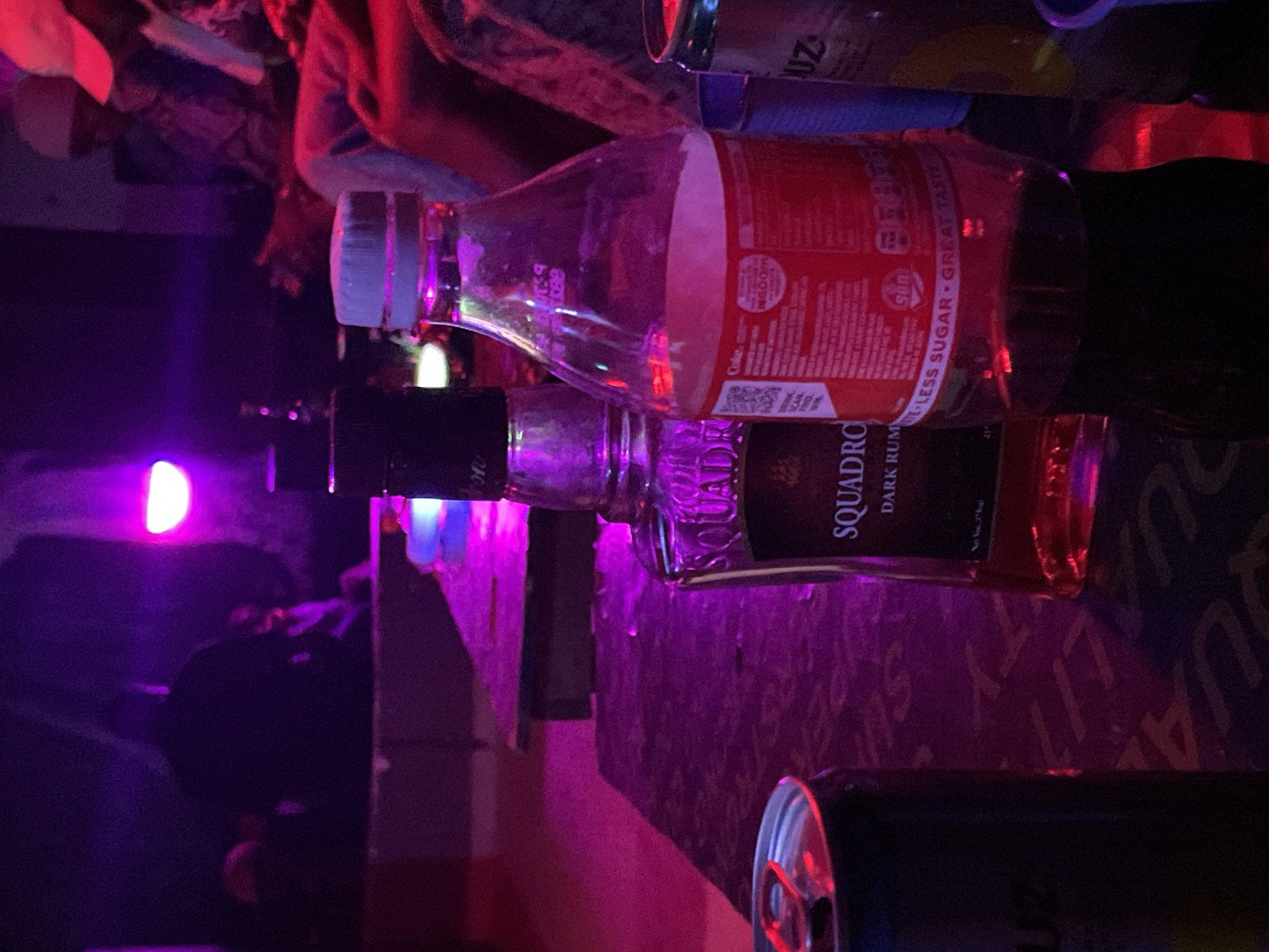 In the city of Durumi, Nigeria, a dimly lit, indoor setting appears to be a lounge or club. The atmosphere is characterized by vibrant purple and red lighting, suggesting a lively or social environment. In the foreground, a dark-colored can, possibly of an energy drink or soda, is visible. Next to it, a bottle of "Squadron Dark Rum" stands upright. To the right, a clear plastic bottle, likely containing Coca-Cola, is also present. The label on the Coca-Cola bottle indicates "LESS SUGAR • GREAT TASTE" and has a QR code. In the background, blurred figures of people are discernible, some seated and others standing, immersed in the ambient light and likely engaged in social interaction. The scene captures a moment of leisure or revelry, with alcoholic beverages and a soft drink readily available.