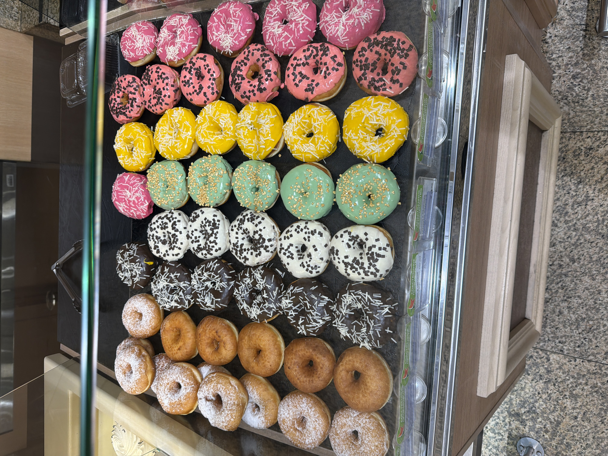 The image shows a display case filled with a variety of donuts. The donuts are arranged in rows, with different colors and toppings. From the top row down, there are donuts with pink frosting and white sprinkles, pink frosting with black sprinkles, yellow frosting with white sprinkles, green frosting with gold sprinkles, white frosting with black sprinkles, brown frosting with white sprinkles, plain donuts, and powdered donuts. The donuts are located in a display case with glass and metal dividers and price tags, positioned against a background of wood paneling and a speckled floor, likely located in a commercial establishment in Yekaterinburg, Russia.