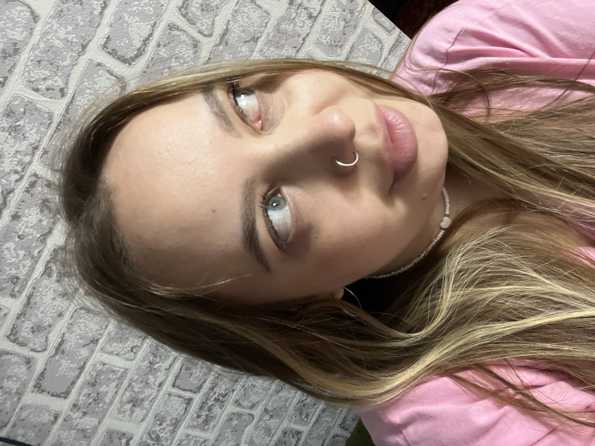 An overhead, tilted indoor portrait captures a young person, likely female, with light brown and blonde-highlighted hair. The subject's head is tilted to the right, and her gaze is directed upwards and to the left, revealing light blue or grey eyes with visible reflections. She has defined eyebrows, plump lips, and a silver nose ring piercing her right nostril.
Her long, wavy hair frames her face and cascades onto her chest, where she is wearing a simple pink top or t-shirt. A delicate white beaded necklace with a small heart-shaped charm adorns her neck. The background consists of a light-colored, textured wall featuring a brick pattern, possibly wallpaper or painted brickwork. The lighting is soft, creating a gentle illumination of her features. No specific activity is discernible beyond posing for the photograph. The expression appears neutral or contemplative. The scene is set indoors, potentially in Novopokrovskaya, Russia. No text is visible in the image.