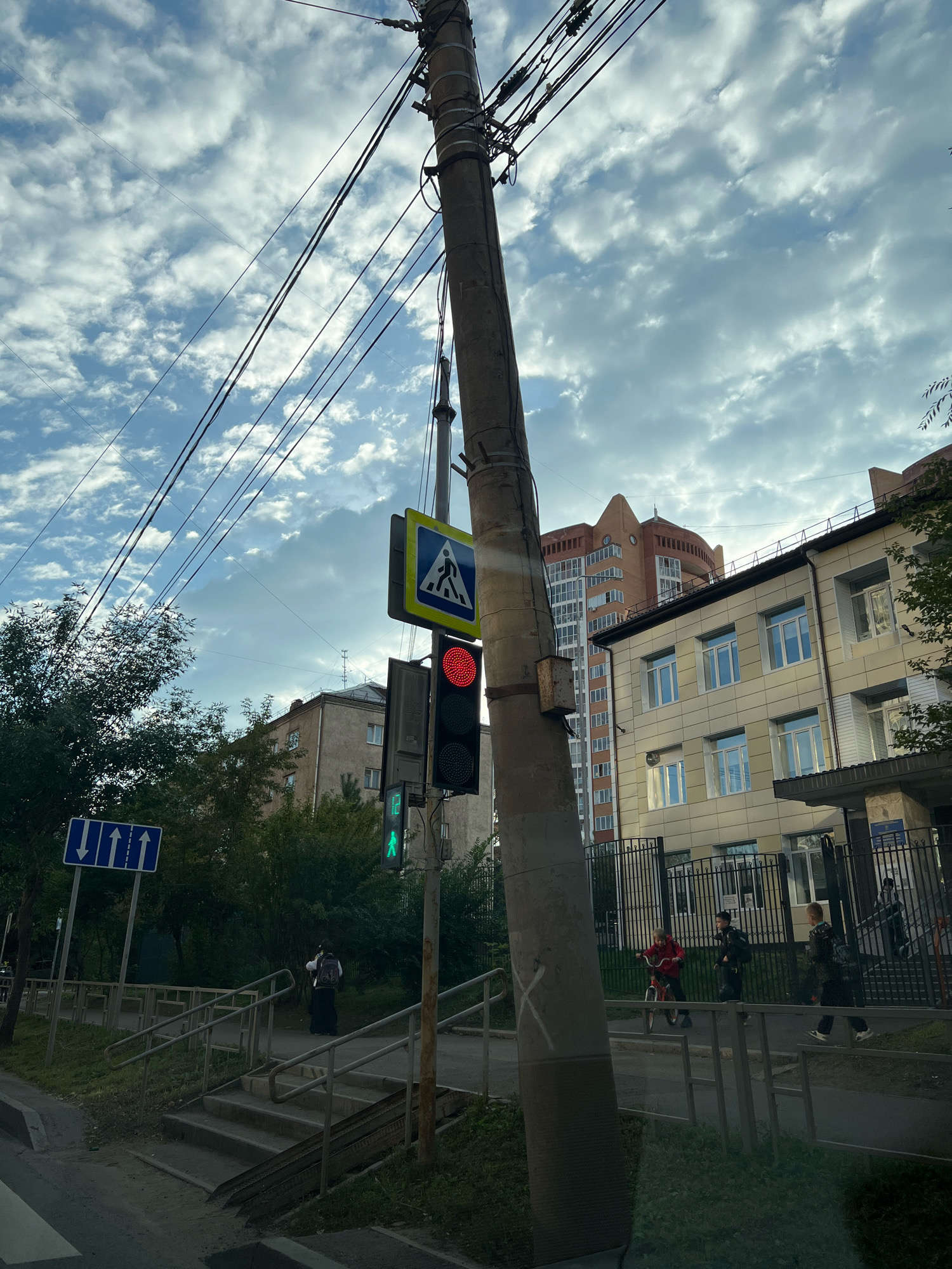 The image captures a street scene in Krasnoyarsk, Russia. In the foreground, a large concrete utility pole dominates the right side of the frame, adorned with electrical wires stretching across the sky, which is filled with a mix of clouds and blue sky. Attached to the pole are traffic signals and signs. The traffic light for vehicles shows a red light, indicating a stop. Above it, a pedestrian crossing sign with a blue background and white pictogram is visible. Below the vehicle traffic light, a pedestrian signal displays a green walking figure, signaling it is safe to cross.
To the left of the utility pole, a blue street sign with white arrows points in the direction of movement. Beyond the signs and traffic lights, a sidewalk with stairs leads up to a street. A person wearing a backpack walks up the stairs.
In the background, several buildings are visible. On the right, a modern light-colored building with multiple windows stands behind a black metal fence. Further behind this building, a taller, multi-story brick building with a distinctive curved top is visible. To the left, there are older, less modern apartment buildings. Trees and greenery line the street, suggesting a residential or mixed-use urban area. The scene appears to be a typical city intersection with pedestrian and vehicular traffic.