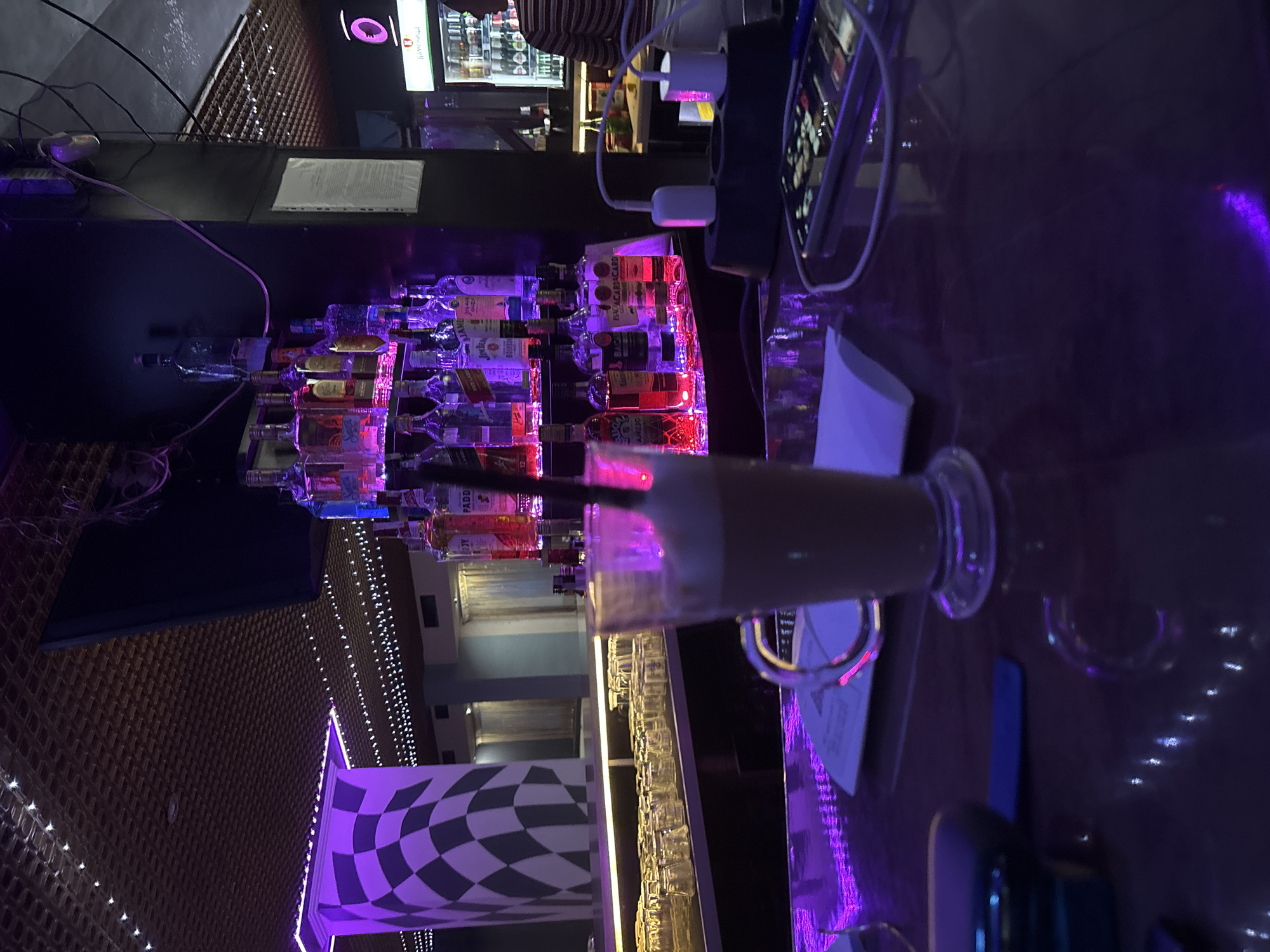 This image captures an indoor scene, likely a bar or club, at night in Chirpan, Bulgaria. The overall atmosphere is dark, illuminated by vibrant purple and red ambient lighting.
In the foreground, a tall glass containing a light brown beverage with a straw rests on a reflective dark bar top. Next to it, a smartphone is connected to a black power strip with white chargers plugged in. A white paper menu or document is also visible on the bar surface.
The midground reveals a well-stocked bar with numerous liquor bottles arranged on tiered shelves, prominently lit by red and purple lights. Brands like "BACARDI" and "PADDY" are discernible on some labels. Below the main bottle display, a row of empty glasses is visible, suggesting a functional bar setup.
In the background, a person wearing a striped shirt is partially visible near a refrigerated display case. Above this display, a small screen or sign shows a purple "IOI" logo and possibly the word "homegood". To the left, a checkered black and white decorative pillar stands out under purple illumination, next to a wall with a textured, possibly woven, pattern that also features scattered white lights. Wires are visible running across a dark surface in the upper left.
The scene depicts a casual evening activity at a lively establishment, with patrons likely enjoying drinks. The artificial lighting, bar setup, and presence of drinks indicate a social, nightlife environment.