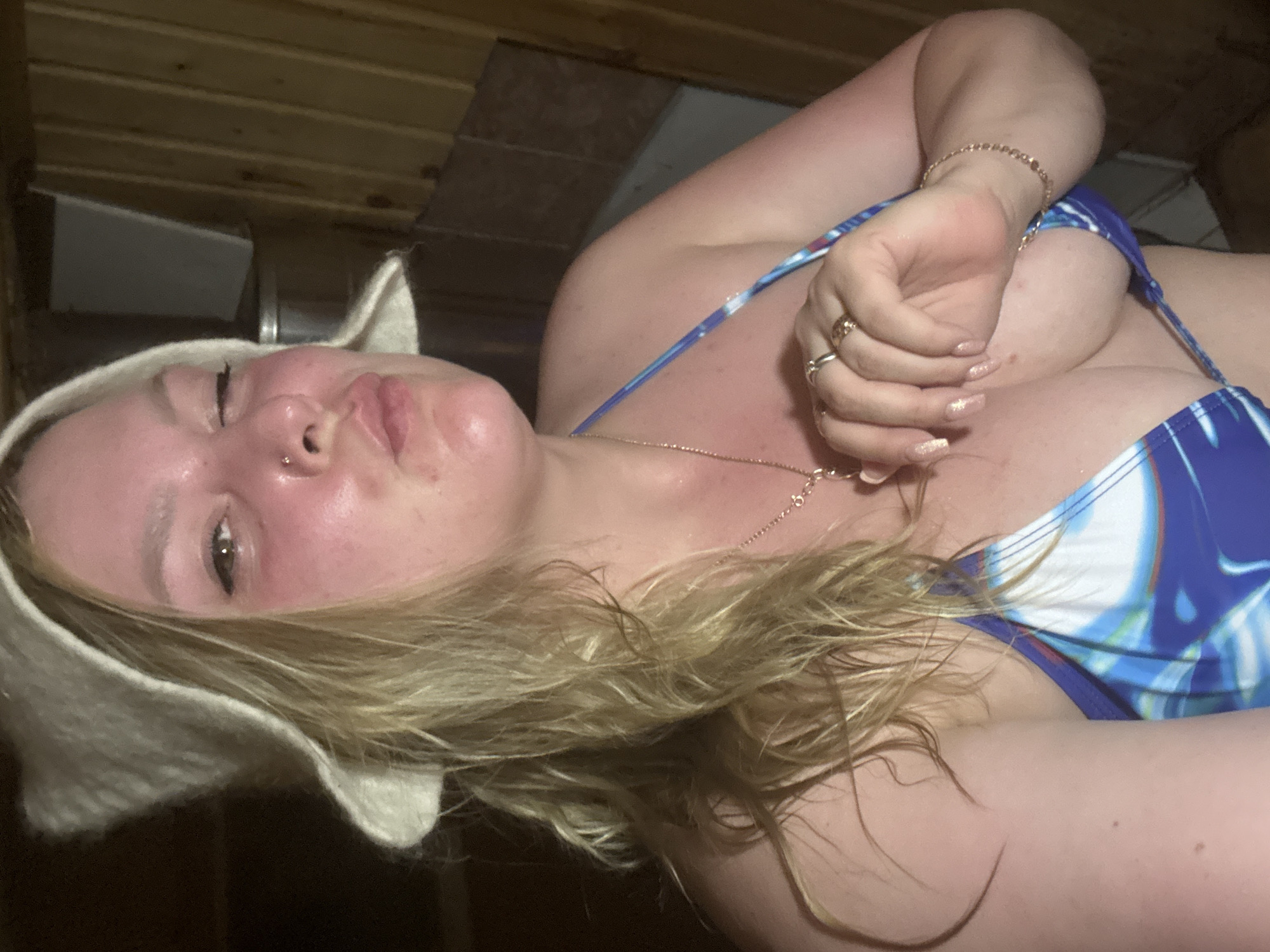 The image shows a woman wearing a white felt hat and a blue and white patterned bathing suit, lying down. She has blonde hair and is wearing jewelry on her fingers, wrist, and neck. The background appears to be a wooden interior, possibly a sauna. There is no indication the photo was taken in the city of Dubovaya, Russia.