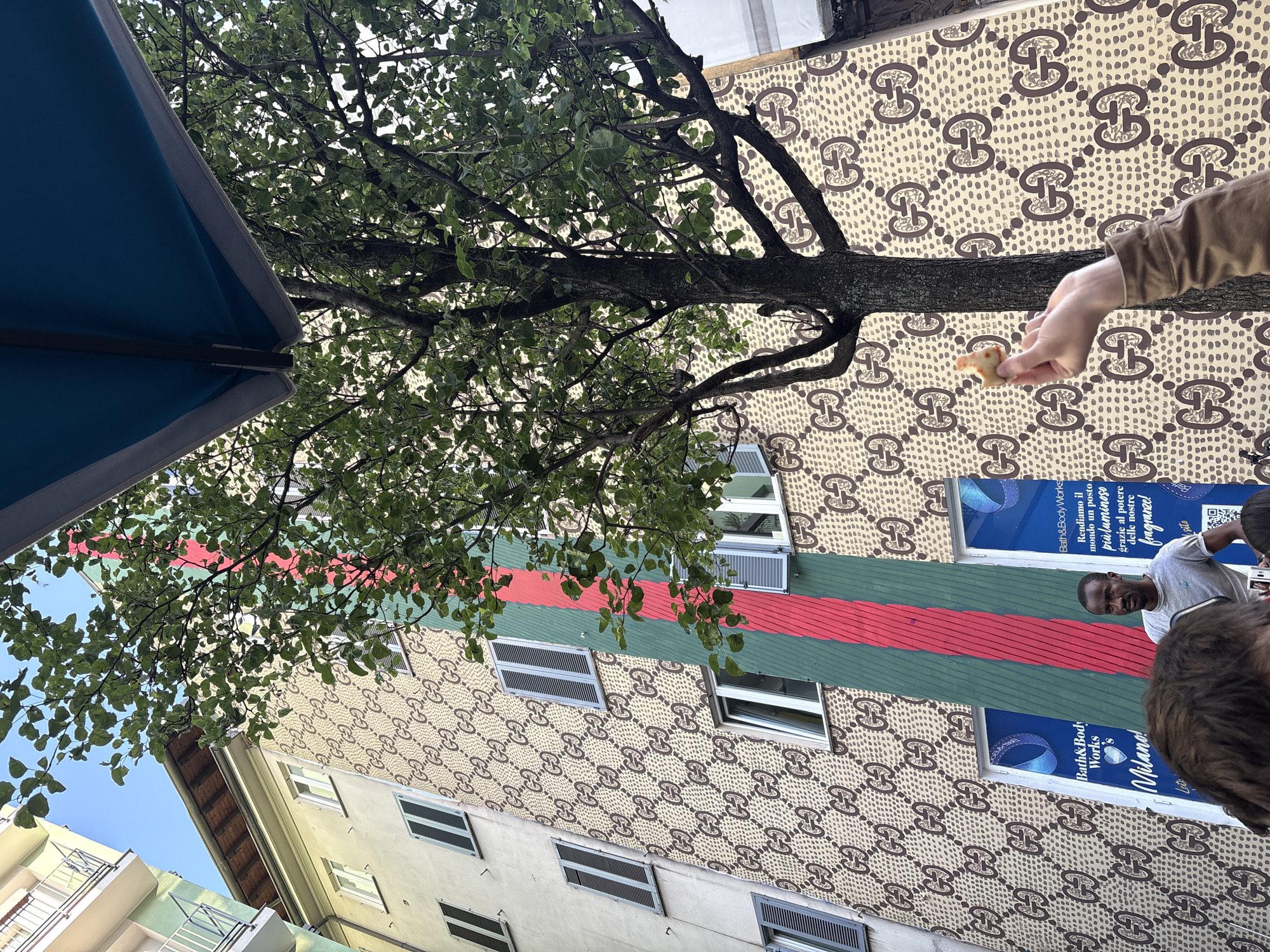 A street scene in Milan, Italy, is captured from a low angle. The facade of a multi-story building is visible, adorned with a repeating pattern reminiscent of the Gucci logo. A green stripe, bordered by red stripes, runs horizontally across the building's exterior. A tree with dense foliage extends its branches across the view. Underneath the tree is a partially visible blue awning. A hand holds a small piece of food in the foreground. A person is partially visible on the right side of the frame, their face visible near a window. The building features advertising posters and several windows with closed blinds.