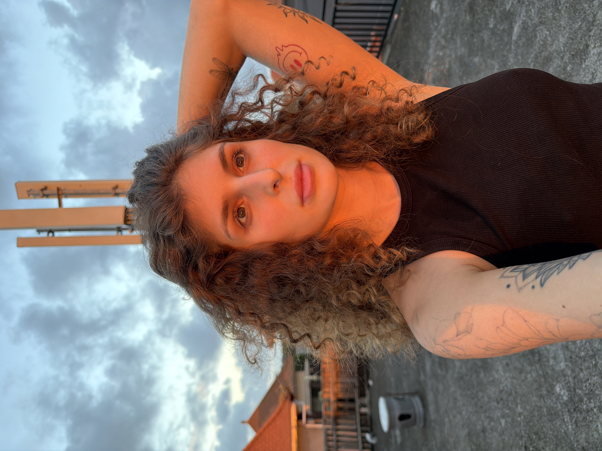 A woman with curly brown hair is standing on a rooftop in North Kuta, Indonesia. She is wearing a black tank top and has tattoos on her arms. The sun is setting, casting a warm glow on her face and the surrounding buildings. The sky is filled with dramatic clouds. In the background, there are tiled roofs of houses and a cell tower.