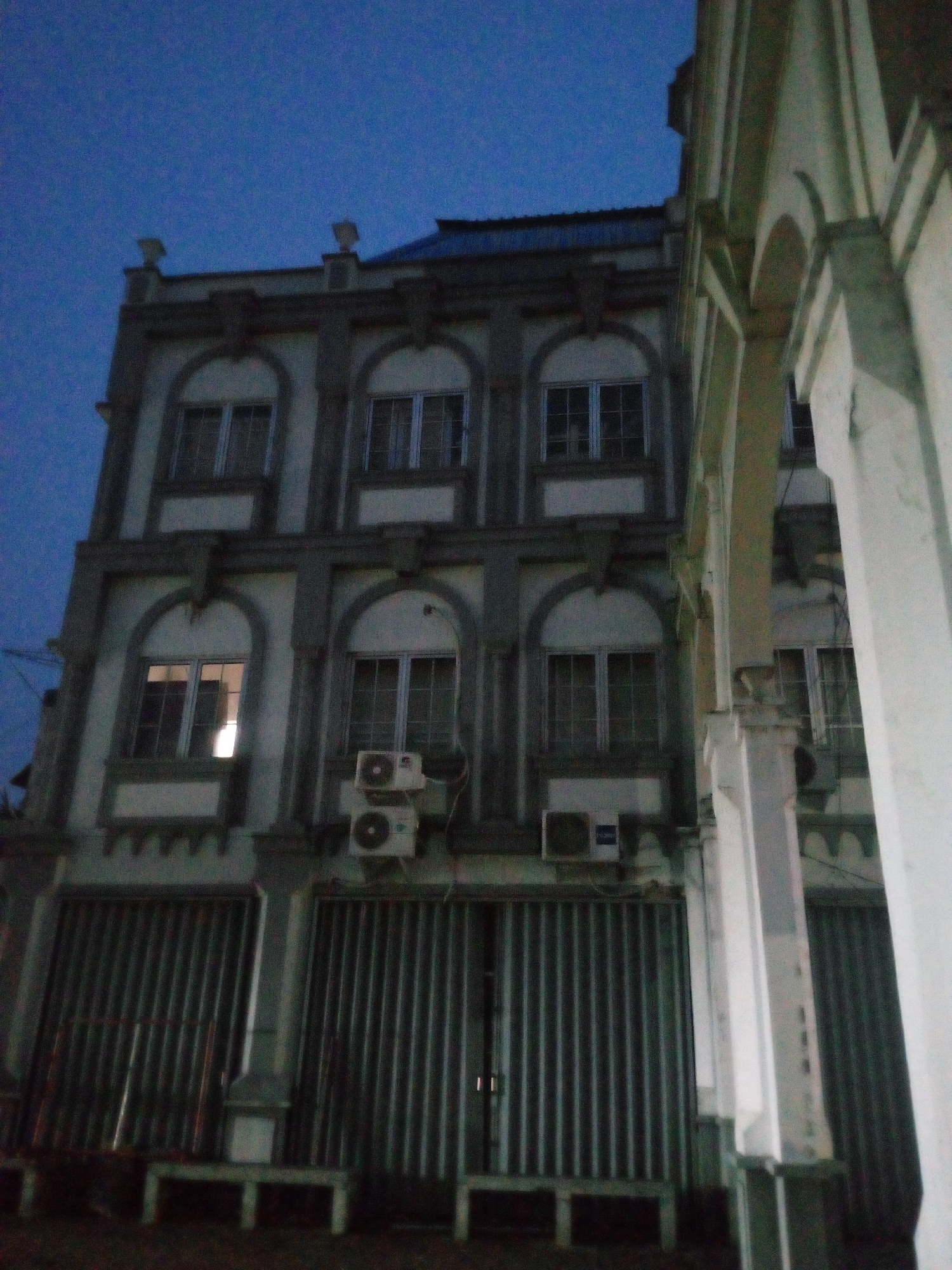 The image depicts a facade of a multi-story building at dusk in Cikupa, Indonesia. The building, likely a commercial or residential structure, exhibits classical architectural elements, including arched windows, decorative molding, and pilasters. Several windows are visible across the different floors, with some appearing illuminated from within. Two air conditioning units are mounted on the exterior wall of the second floor. The ground level features large, corrugated metal shutters. To the right of the building, a portion of another structure with arched openings and substantial white columns is partially visible, suggesting a connected or adjacent building with a similar architectural style. The sky is a deep blue, indicating twilight. No people or vehicles are discernible in the scene. The environment appears to be an urban setting.