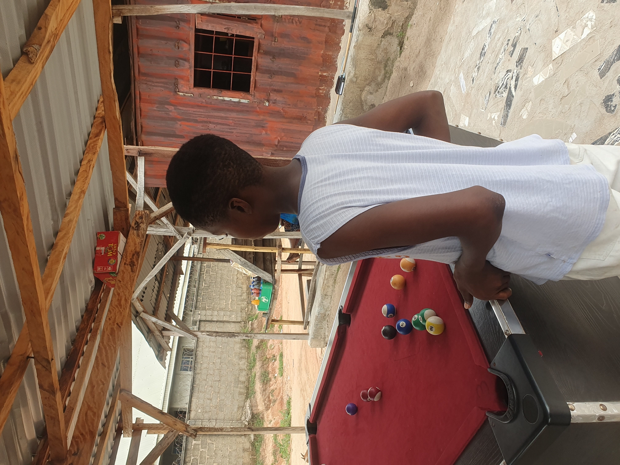 A young person with short, dark hair stands facing away from the camera, looking towards a red billiard table. They are wearing a sleeveless white t-shirt and white pants. The billiard table has several balls scattered across its surface, including a purple ball, a striped red and white ball, a black ball with a blue number 10, a blue ball with a white number 2, a green ball with a white number 3, a yellow ball with a white number 9, a striped yellow and white ball, and a striped orange and white ball. The table is situated outdoors under a wooden and corrugated metal roof structure. In the background, there is a red corrugated metal building with a window grid and a low brick wall. The ground is a mix of dirt and paved areas with some debris. To the left of the billiard table, a wooden stand holds several bottles of beverages. The scene appears to be in Lokoja, Nigeria, possibly a recreational area or a casual establishment.