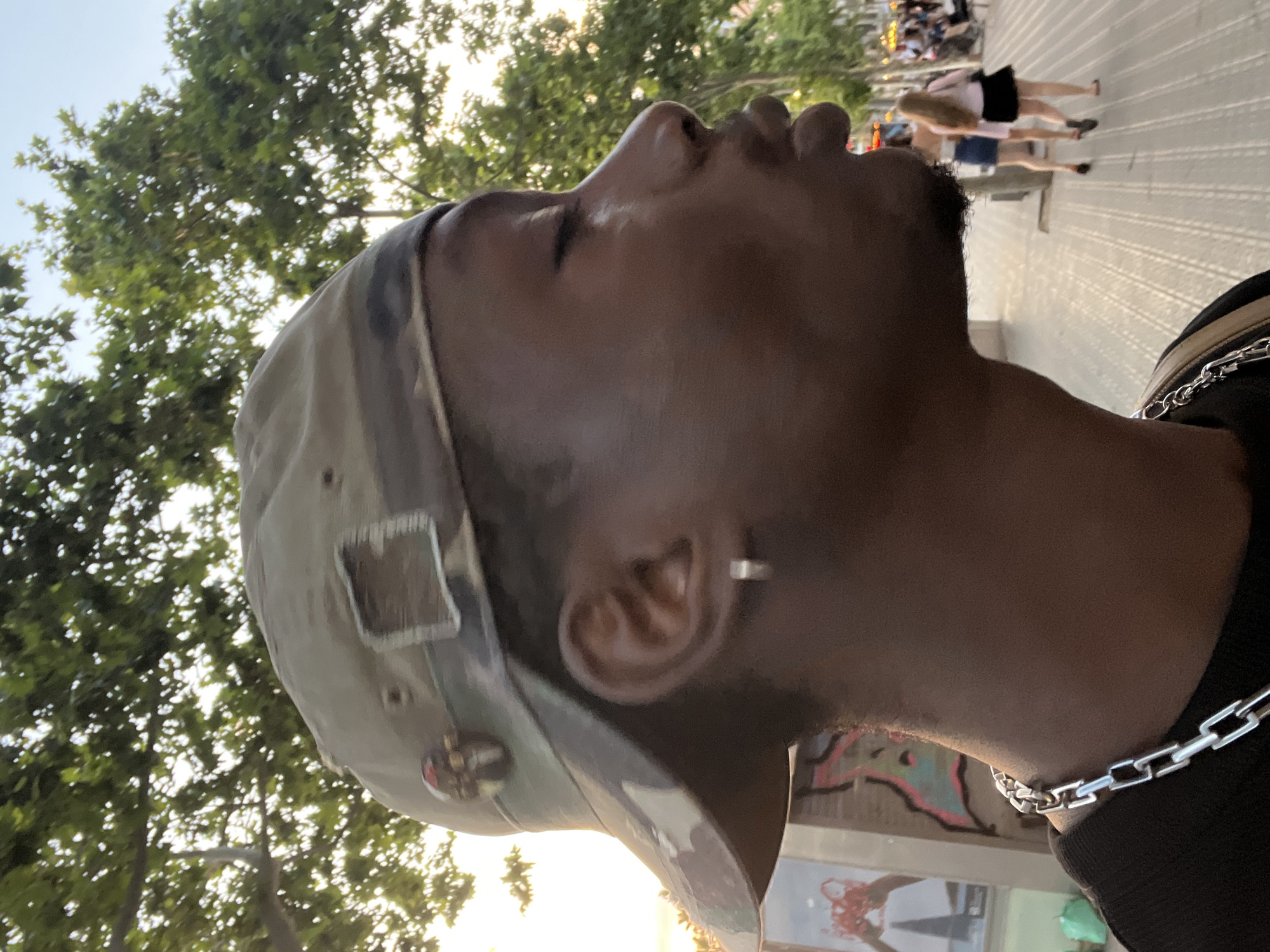 This is a medium shot, profile view photograph of a young Black man wearing a camouflage baseball cap and a silver chain necklace. He is outdoors with trees and a clear sky visible in the background. The man's eyes are closed, and his lips are slightly pursed. He is wearing a silver hoop earring in his left ear. The background suggests an urban environment, possibly a pedestrian walkway, with other people visible in the distance. The lighting indicates it may be late afternoon or early evening. The image was taken on La Rambla in Barcelona, Spain.