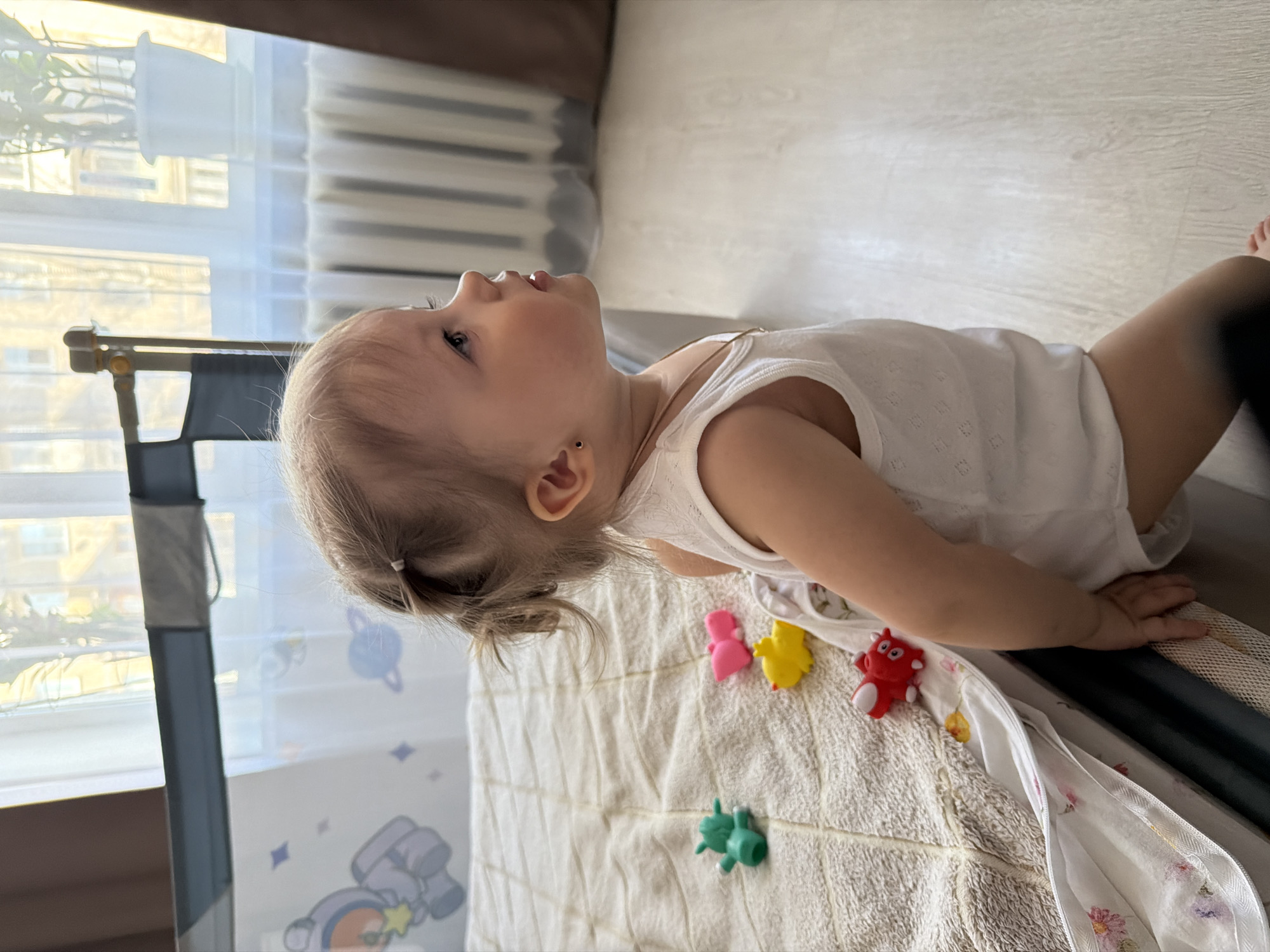 A young child is depicted standing in a room in Reshetnikovo, Russia, observed in profile facing right. The child has fair skin, light-colored hair tied into two small ponytails, and wears a white, sleeveless, patterned bodysuit. A small dark stud earring is present in the visible earlobe, and a thin gold-colored necklace is around the neck. The child's left hand is placed on the edge of a bed or playpen, which features a light-colored quilted blanket and a white side panel decorated with purple astronaut, planet, and star motifs. Four small, colorful plastic animal toys (green, pink, yellow, and red) are scattered on the blanket. In the background, a large window is visible, covered by a sheer white curtain. Through the window, parts of light-colored buildings are discernible under natural daylight. A white potted plant sits on the windowsill to the right. Below the window is a white, finned radiator. The floor appears to be light-colored laminate or wood. A dark brown curtain or furniture edge is present on the far right of the frame.