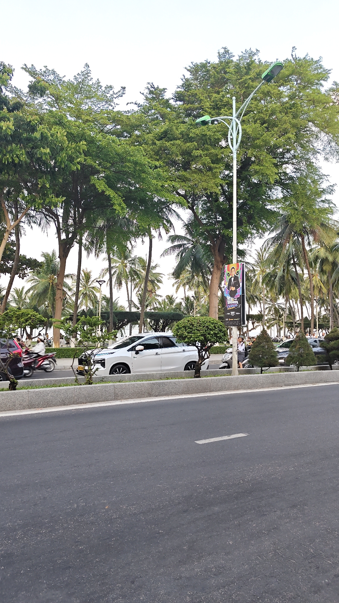 The image depicts a street scene in Nha Trang Ward, Vietnam, characterized by a lush green environment bordering a roadway. The foreground is dominated by a paved road with white lane markings, suggesting a public thoroughfare. To the left of the road, a grey concrete divider separates it from a sidewalk or median strip. Along this strip, several well-maintained trees and manicured shrubs are visible.
In the middle ground, a white sedan is parked or driving slowly on the road. Further back, palm trees and other deciduous trees form a dense canopy, creating a tropical atmosphere. A street lamp with a curved design and two green lampshades stands prominently on the right side of the image, with a colorful banner advertising an event attached to its pole. The banner features a man in a tuxedo and text in what appears to be Vietnamese.
Several motorcycles are also present on the road, with one rider in the middle left section of the image wearing a white helmet and a dark jacket. A person in a uniform, possibly a security guard or attendant, is visible near a parked scooter on the right side, standing next to the street lamp. The background is largely obscured by the dense foliage, but a glimpse of buildings and more trees suggests an urban or suburban setting. The overall lighting suggests it is daytime, possibly late afternoon, with the sky appearing overcast or bright white.