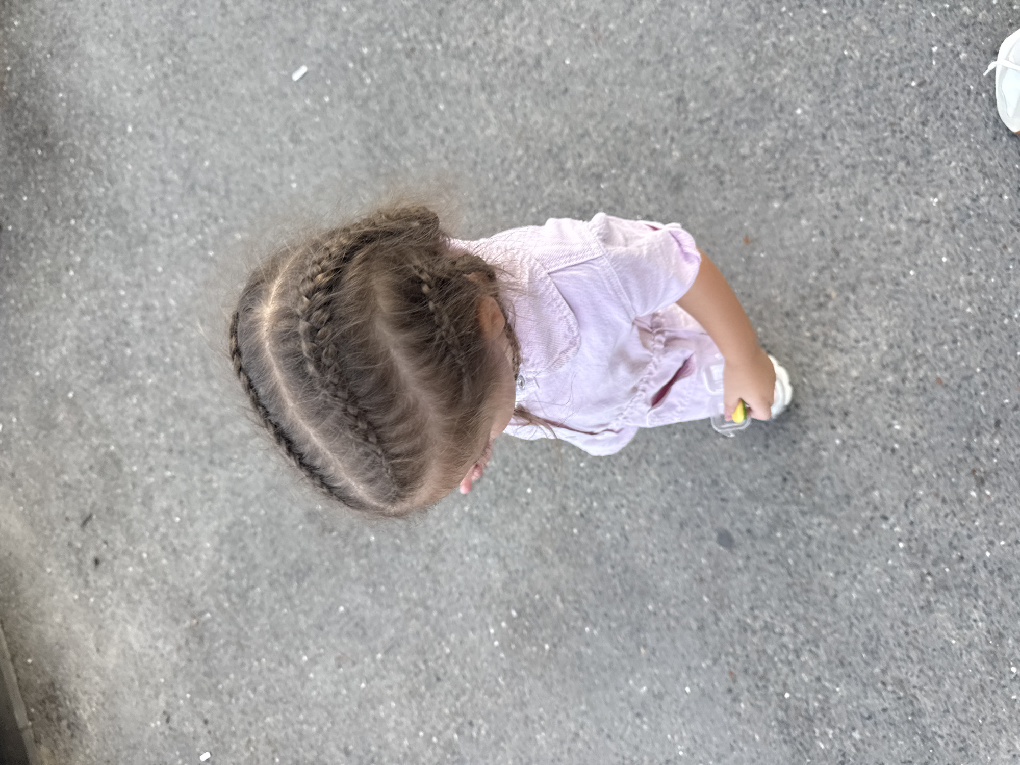 An overhead shot shows a young child from the shoulders up, facing away from the camera and slightly to the left. The child has braided dark hair, styled into two prominent braids that run from the forehead towards the back of the head. The child is wearing a light pink, collared shirt with some sort of detailing or pattern on the fabric, possibly a textured or embroidered design. In the child's right hand, a small, bright yellow object is visible, held within a clear container. The child's left hand is partially obscuring their face. The background is a gray asphalt surface, appearing to be a street or pathway, with scattered small white and dark particles. The lighting suggests it is daytime. The image does not contain any identifying features or landmarks that would allow for specific location identification within Chișinău, Moldova.