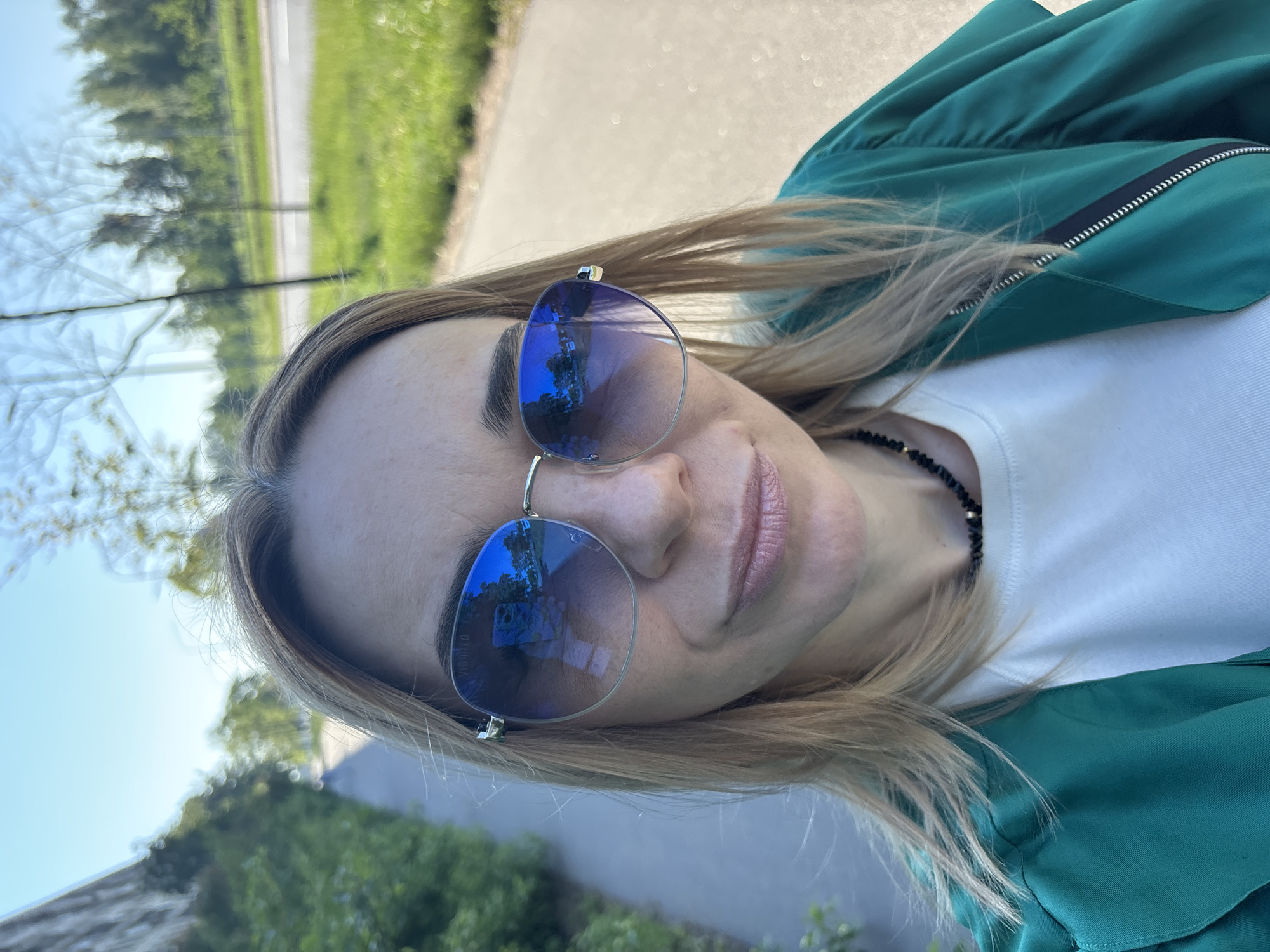 An outdoor portrait shows a person, likely a woman, looking towards the viewer. The image is taken at a slight upward angle and is rotated 90 degrees clockwise. The subject has long, light-colored hair that flows over her shoulders and jacket. She is wearing large, blue-mirrored aviator-style sunglasses that reflect trees and what appears to be a mobile phone or camera. A faint brand name, "VIGNATO" or "VIORATO," is visible on the sunglasses lens.
She is dressed in a vibrant green jacket with a black and white striped zipper detail on the front, worn over a white t-shirt. A black beaded necklace with a small silver bead is visible around her neck. Her lips are covered in a light pink lipstick, and her facial expression appears neutral or slightly smiling. Some minor skin imperfections or freckles are visible on her forehead and nose.
The background, partially blurred, indicates an outdoor setting with ample sunlight. Green foliage, trees, and a paved road or path can be seen behind her, suggesting a park, a walking trail, or a street. The bright lighting indicates it is daytime with clear weather. The overall impression is a casual outdoor shot, possibly a selfie or a candid moment.