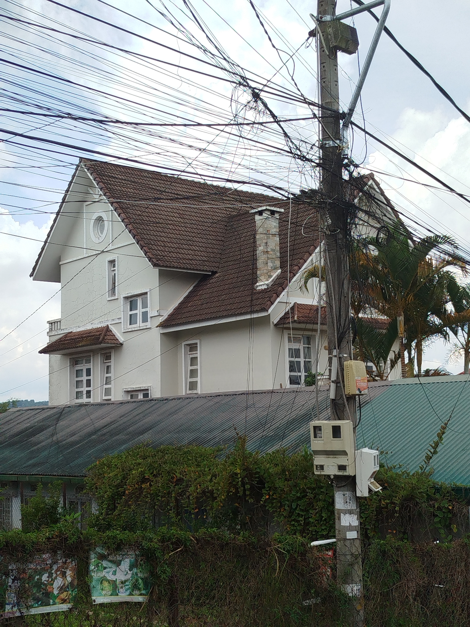 The image depicts a view of a residential building in Lâm Viên - Đà Lạt, Vietnam, on an overcast day. A prominent utility pole with a dense entanglement of wires dominates the foreground, obscuring parts of the building. The building itself is light-colored with a brown tiled roof and features multiple windows and a chimney. In front of the building, a corrugated metal roof covers another structure, and dense green foliage with some posters is visible at the bottom of the frame. The overall scene suggests a somewhat cluttered urban environment with a mix of residential architecture and essential infrastructure.