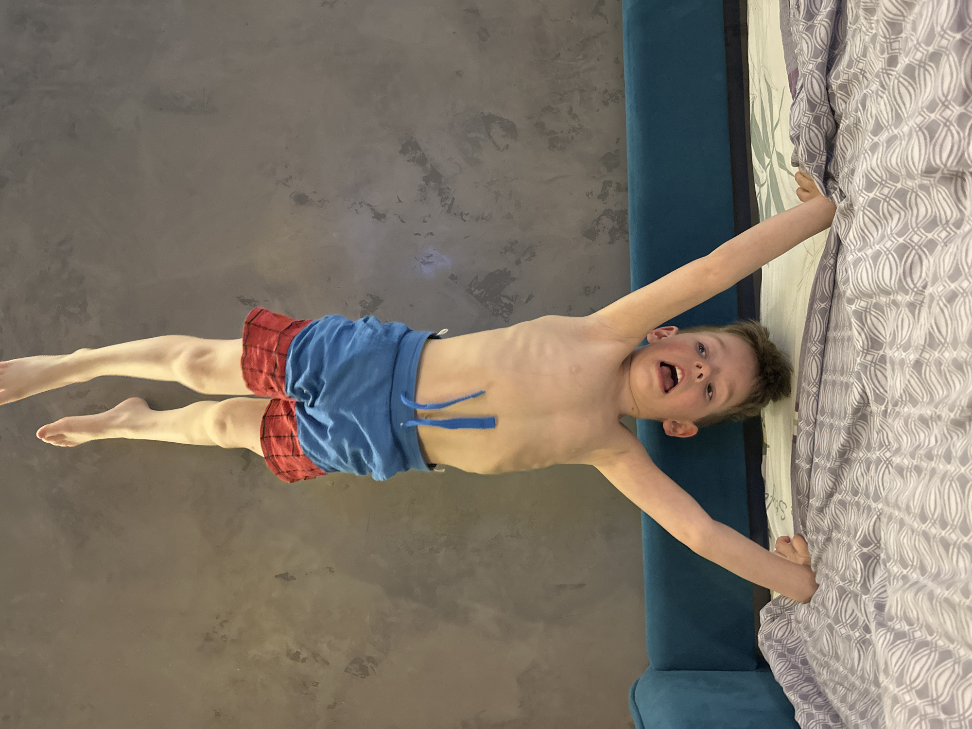 A young, shirtless boy is lying on his back on a gray floor, with his arms stretched upward toward a blue sofa and a patterned bedspread. He is wearing blue shorts with a red and black checkered pattern at the hem. The boy is Caucasian, has short brown hair, and is looking toward the viewer with an open mouth. The sofa is teal in color. The bedspread has a gray and white geometric pattern. The location cannot be confirmed as Прикамский, Russia from the context given.