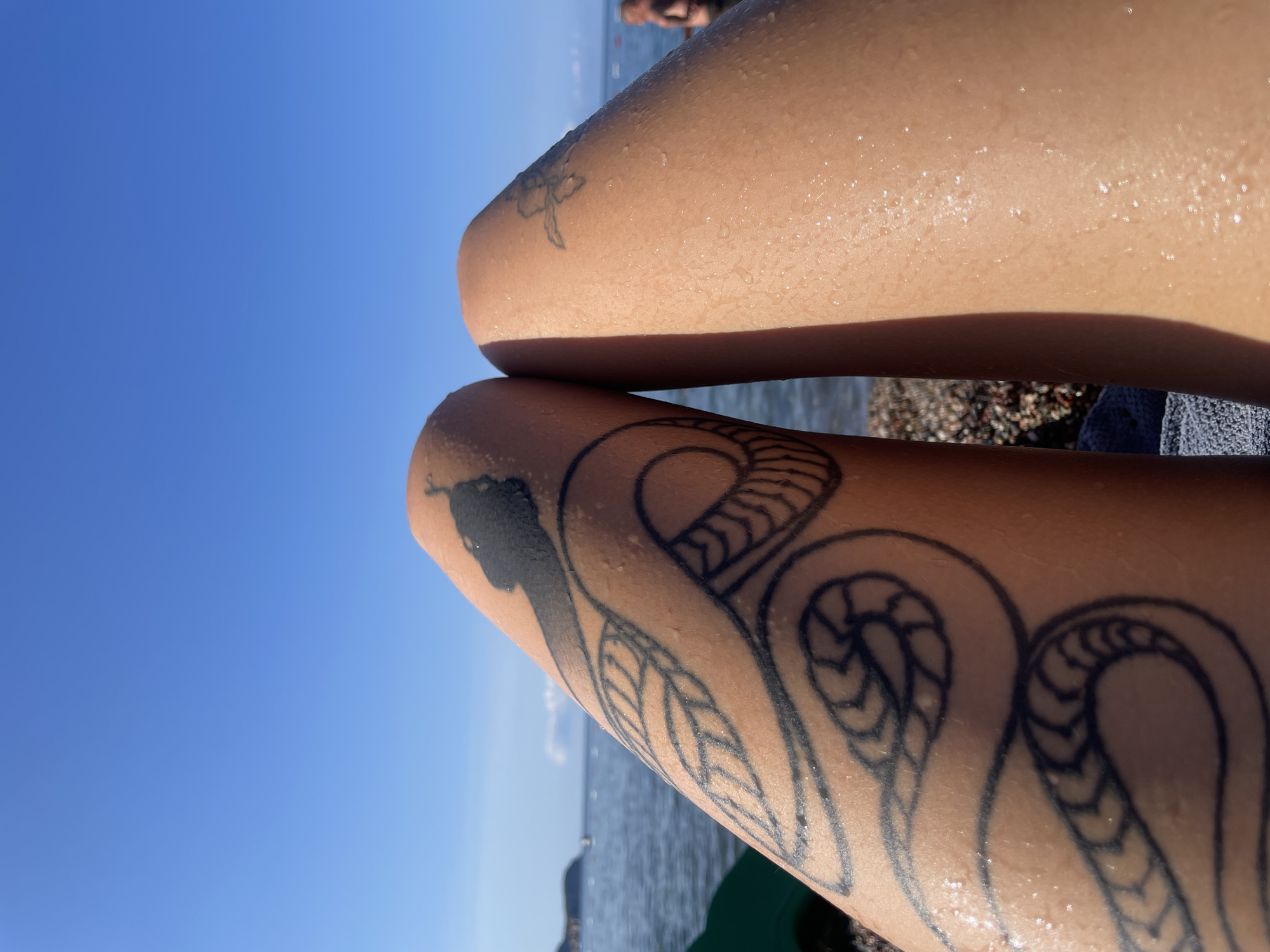 In Rafailovići, Montenegro, on a bright, sunny day, the image captures a close-up perspective of a person's legs, resting near the water's edge. The legs are tanned and slightly damp, with a prominent black snake tattoo coiled around the left thigh, extending towards the knee. Another smaller, faded tattoo, possibly a floral design, is visible on the right thigh. The background reveals the serene Adriatic Sea, with a clear blue sky overhead. Distant boats can be seen on the water, and a blurred figure of another person is visible further down the beach. The foreground consists of a pebbled shoreline and a glimpse of a green object, potentially beach equipment. The overall scene evokes a sense of relaxation and enjoyment of a day at the beach.