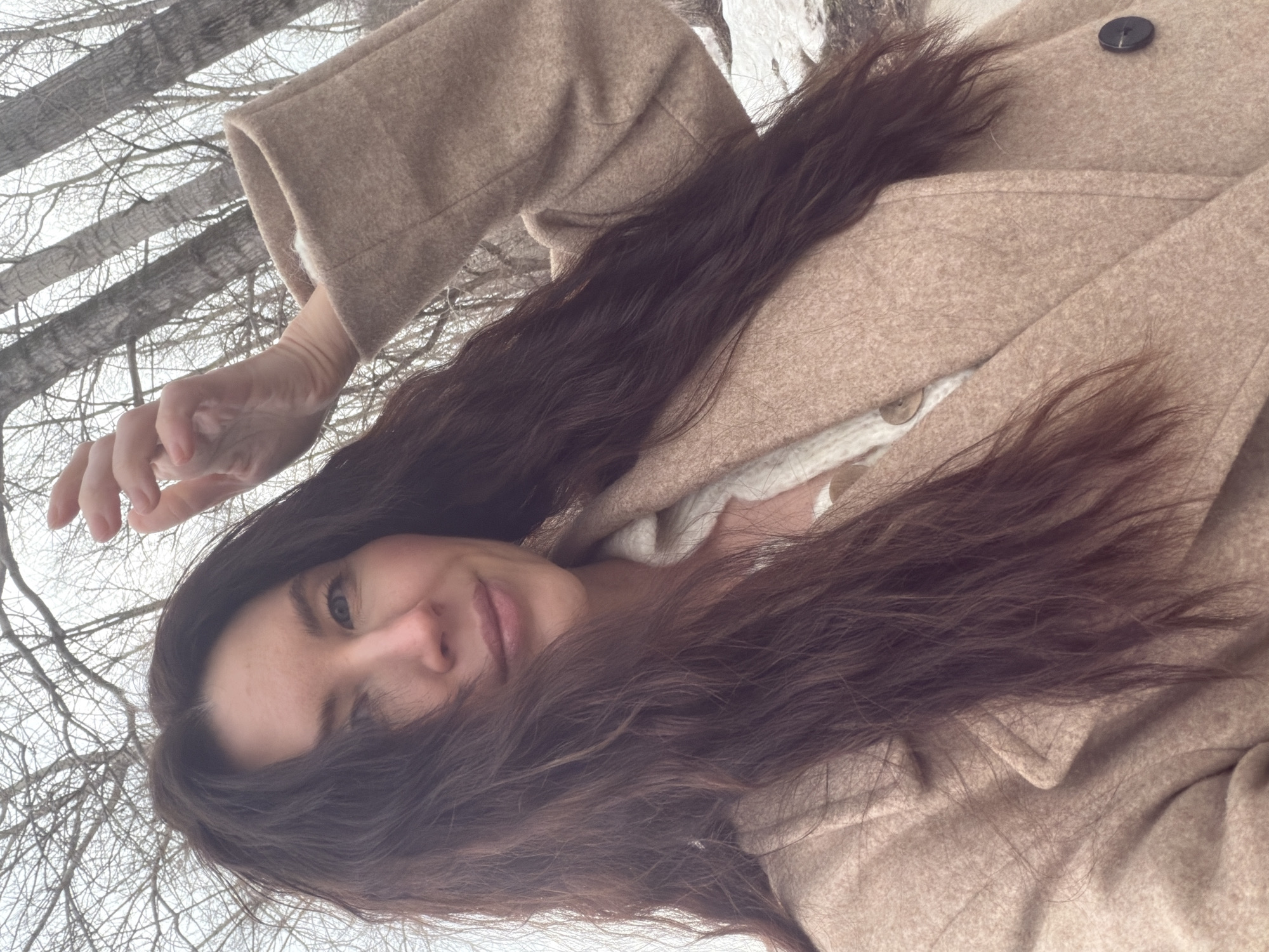 An adult female is depicted in a selfie taken outdoors in Perm, Russia. She has long, dark brown hair and blue eyes, displaying a subtle smile as she looks directly at the camera. She wears a light beige wool coat over a cream-colored, buttoned knitted top. Her right hand is raised above her head, with fingers slightly curled. The background consists of numerous bare deciduous trees, indicating a season of defoliation such as late autumn, winter, or early spring. Patches of snow are visible on the ground, and the sky is overcast.