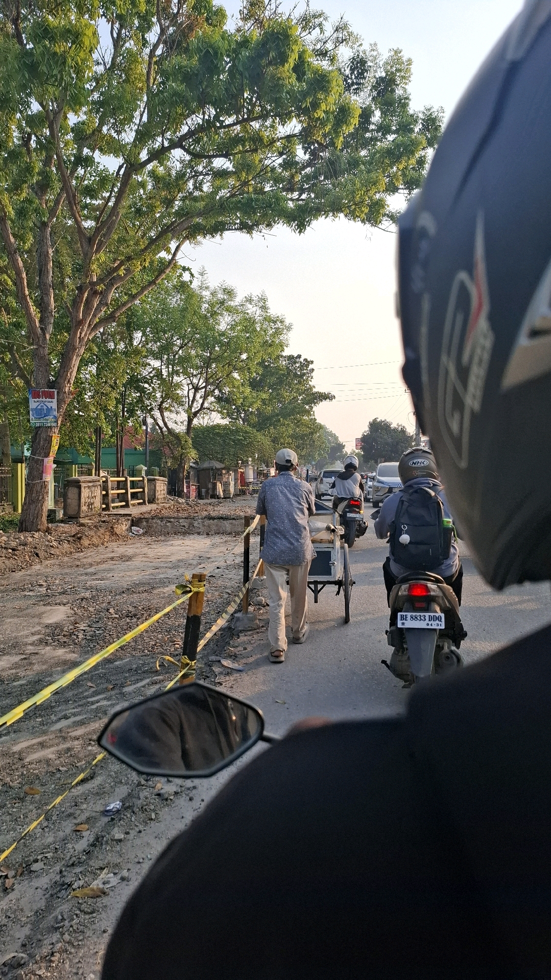 This is a first-person perspective photograph taken from a motorcycle, looking forward and slightly to the left. The scene appears to be a street in Bandar Lampung, Indonesia, undergoing construction or repair. In the foreground, the back of a black motorcycle helmet is visible, along with a portion of the rider's black jacket and a side mirror.
The road surface is a mix of asphalt and gravel, with yellow caution tape marking off a section of the road. A man wearing a light-colored collared shirt, beige pants, and a white cap is seen walking away from the camera, pushing a handcart. He is on the left side of the road, beside the construction area.
Further ahead on the road, a motorcycle with a rider wearing a backpack and a helmet with "NHO" branding is moving away from the camera. Another motorcycle is visible in front of it, with its rider also wearing a helmet. Several cars are also in motion in the background, indicating moderate traffic.
Large trees with green foliage line the left side of the road, casting shadows on the street. In the background, several small buildings and makeshift stalls are visible. The sky is bright, suggesting it is daytime, possibly late afternoon due to the quality of light. The overall impression is a typical urban street scene with ongoing infrastructure work.