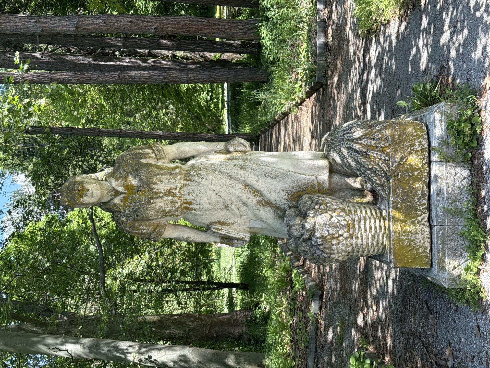 The image shows a weathered stone statue of a woman in a park setting, likely located in Pleven, Bulgaria. The statue, partially covered in moss or lichen, stands on a stone base alongside a walking path. The woman is depicted with drapery and holding or leaning on what appears to be a basket or some other object. The scene is outdoor, with trees in the background and dappled sunlight filtering through the canopy. The walking path is paved with gravel.