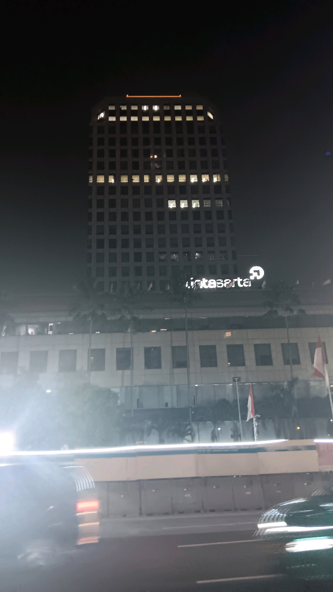 The image depicts the "Inti Jakarta" building at night. The building is tall and features numerous windows, many of which are illuminated. A sign with the text "inti sarta" and a logo is visible on the side of the building. In the foreground, blurry motion of vehicles on a road is apparent, suggesting the photo was taken from a moving vehicle. Two Indonesian flags are also visible on poles on the lower section of the building. The scene is illuminated by artificial lights, and the overall atmosphere is one of a city at night.